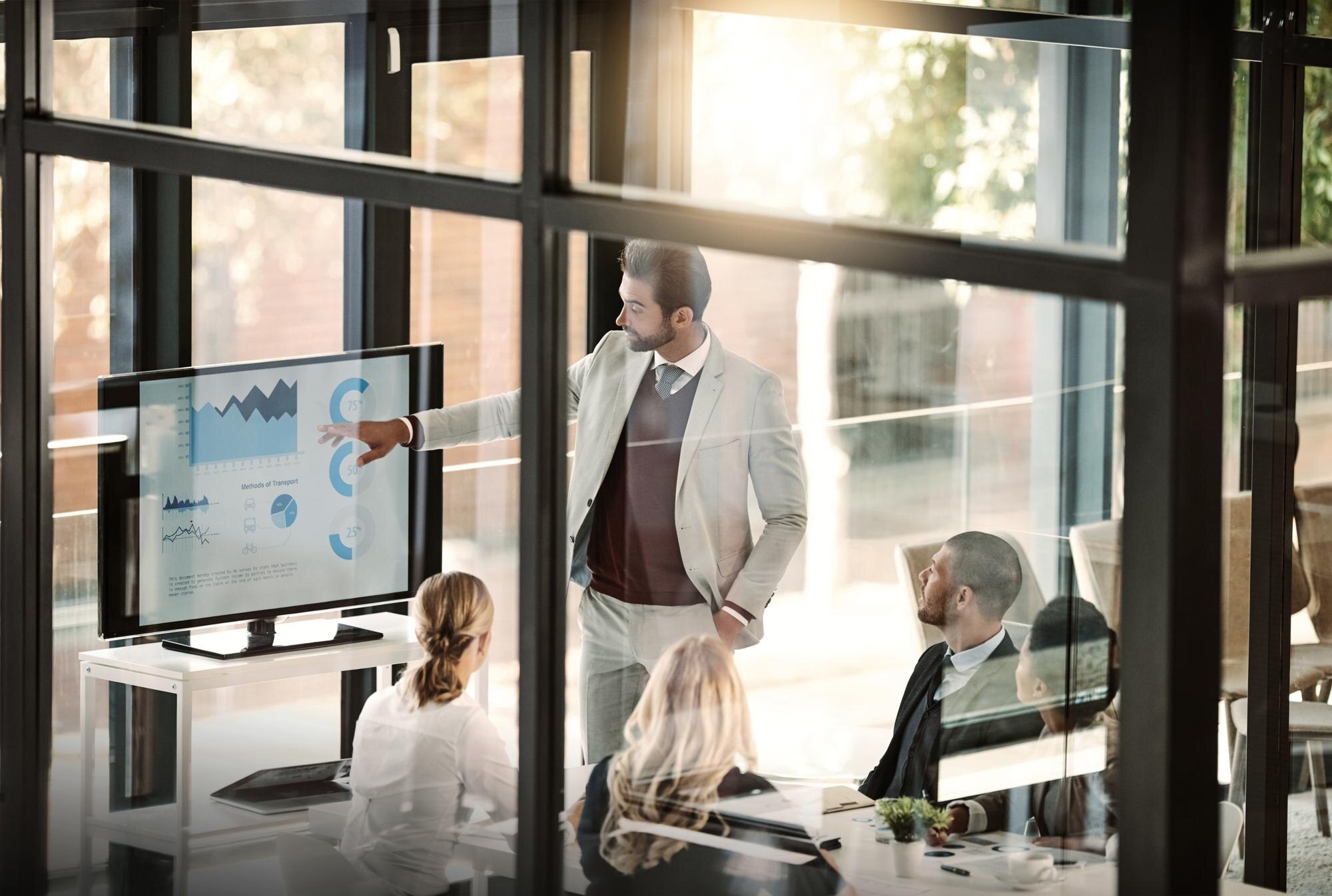 Someone presents data from a large meeting room display while a group of individuals sit at the table, listening. 