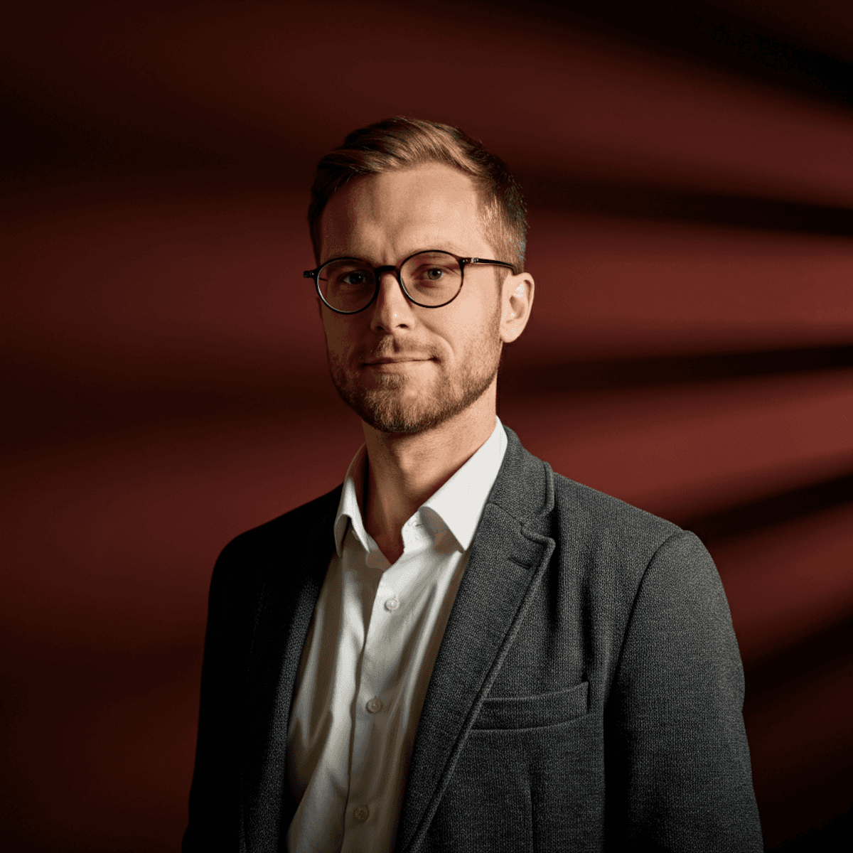 A man with glasses and beard, in a gray blazer and white shirt, stands confidently against a dramatic, dark red background with shadowed lines.