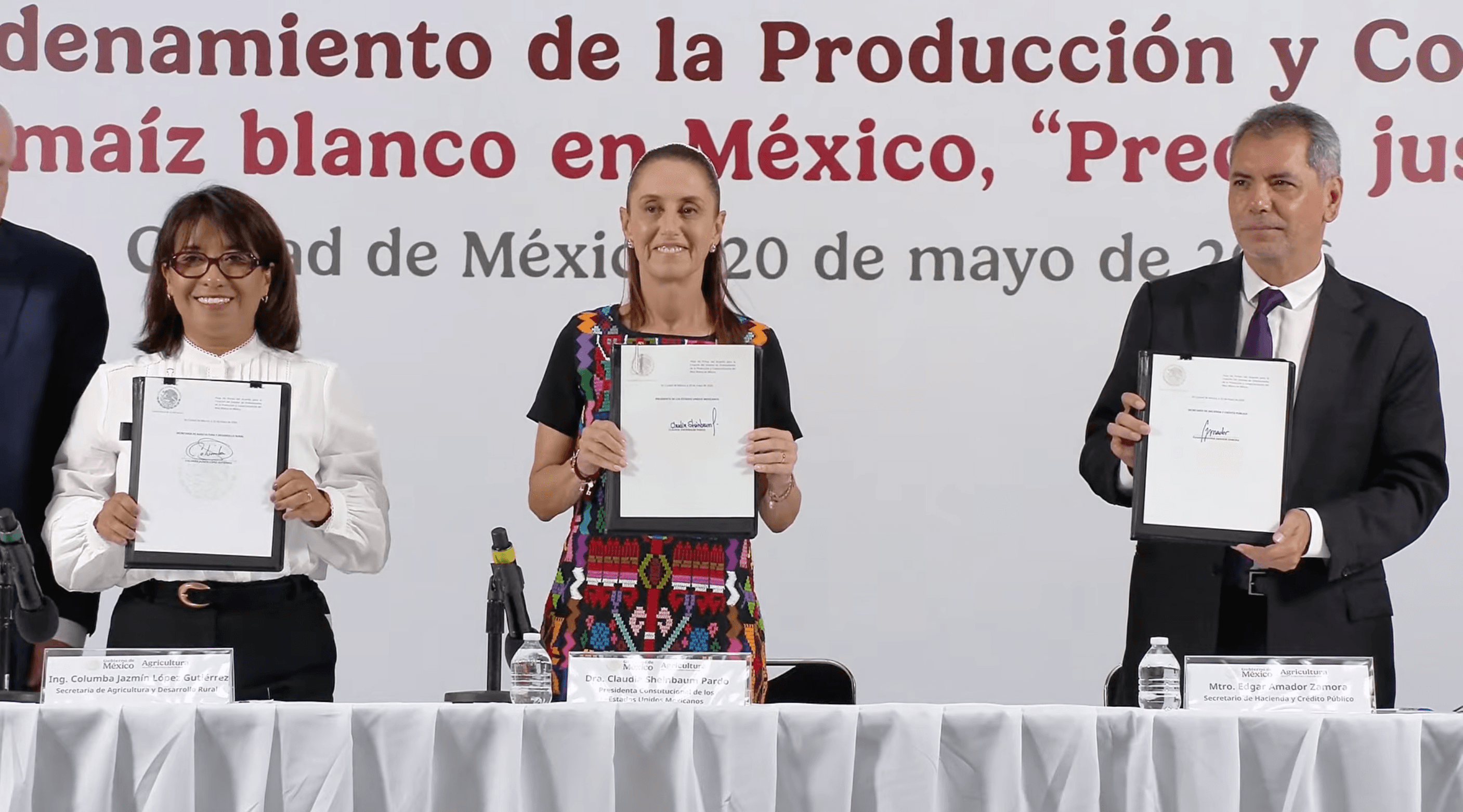 Claudia Sheinbaum and two officials smile while standing behind a table and holding up signed official documents at a Mexican government event regarding white corn production.