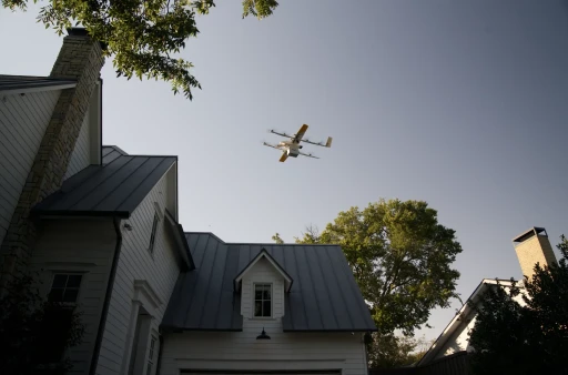 A drone with multiple propellers flies over a suburban neighborhood, casting a shadow on a house with a gray metal roof and surrounded by lush green trees under a clear blue sky.