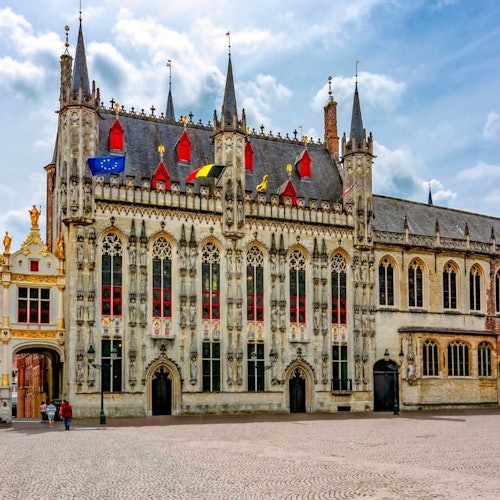 Građevina u gotičkom stilu s raskošnim detaljima, tornjevima i zastavama. Susjedni lučni prolaz. Trg popločan kamenom u prvom planu. Ljudi na lijevoj strani.