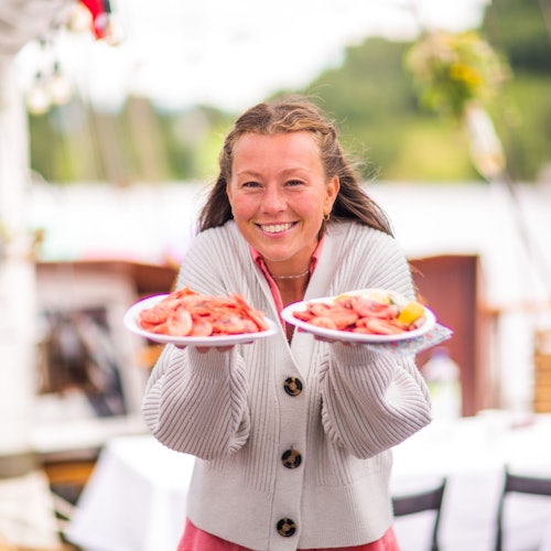 Улыбающийся человек в сером свитере держит две тарелки с креветками, на фоне открытого пространства с природным окружением.