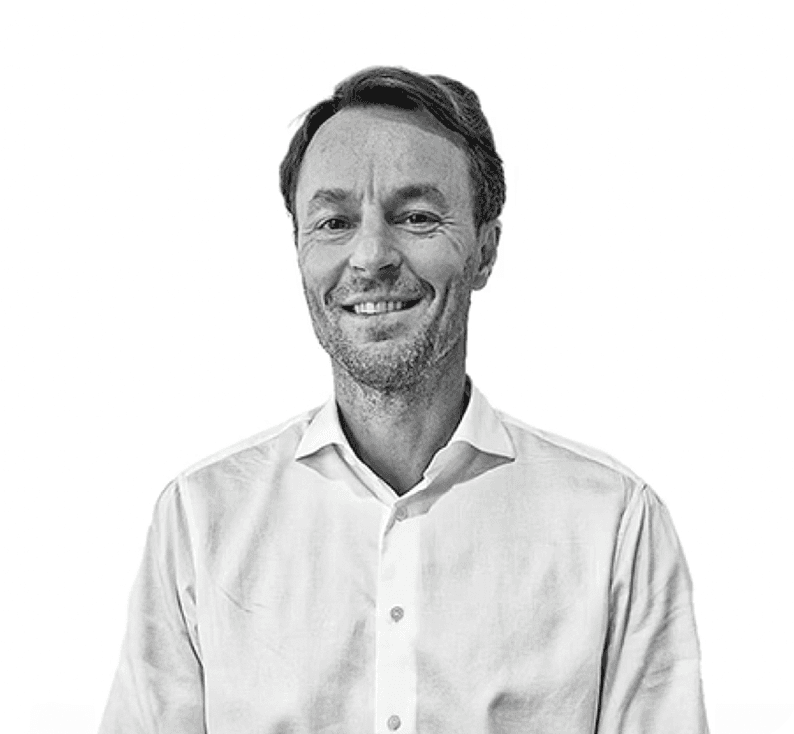 A focused woman with long red hair is seated at a desk, wearing a black blazer and dark blouse. She intently looks at camera, smiling slightly, with an open laptop in front of her. The setting is a warm and serene office setup in the background. Soft daylight filters through, creating a calm atmosphere. The composition captures her in a frontal profile with a clear emphasis on her concentration and the serene indoor setting.