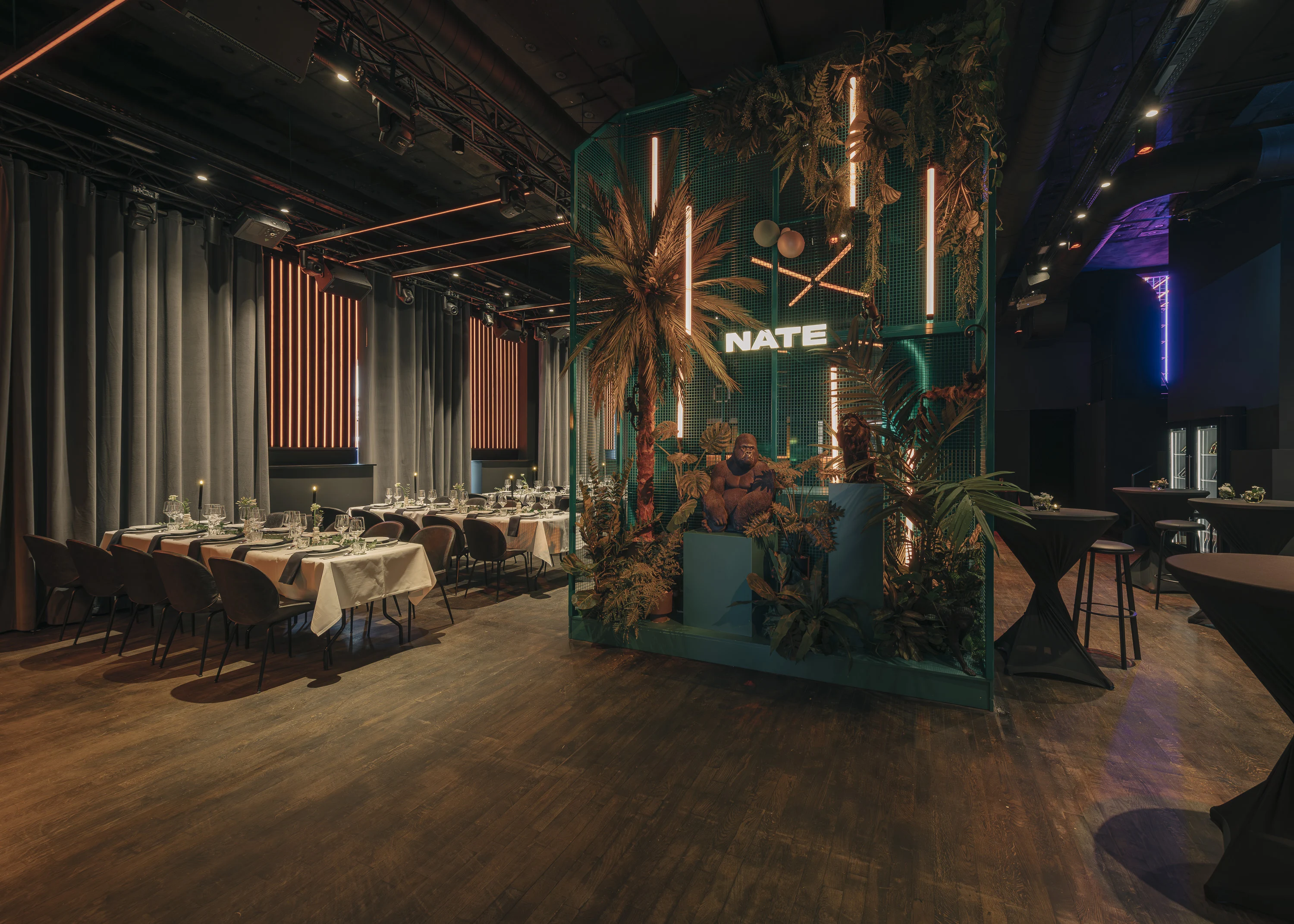 Event hall at NATE CLUB with long white-clothed dinner tables, a teal decor wall with palm plants, neon tubes and an illuminated NATE logo.