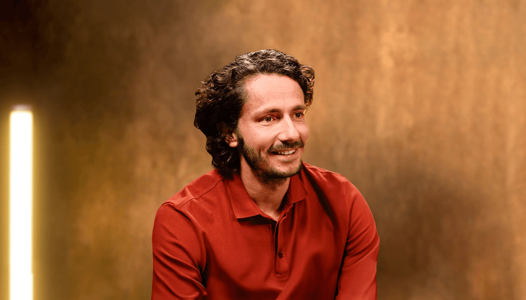 Portrait professionnel et souriant de Guillaume Pley, fondateur du média digital Legend, dans son studio d'enregistrement. Il est vêtu d'un polo rouge terre cuite. Le sujet regarde vers la droite, laissant un large espace négatif à droite de l'image. L'arrière-plan est un mur texturé, flou, dans des tons chauds (or, bronze, marron), éclairé par une colonne lumineuse verticale à l'extrême gauche, créant une ambiance de studio d'interview et de podcast haut de gamme. 