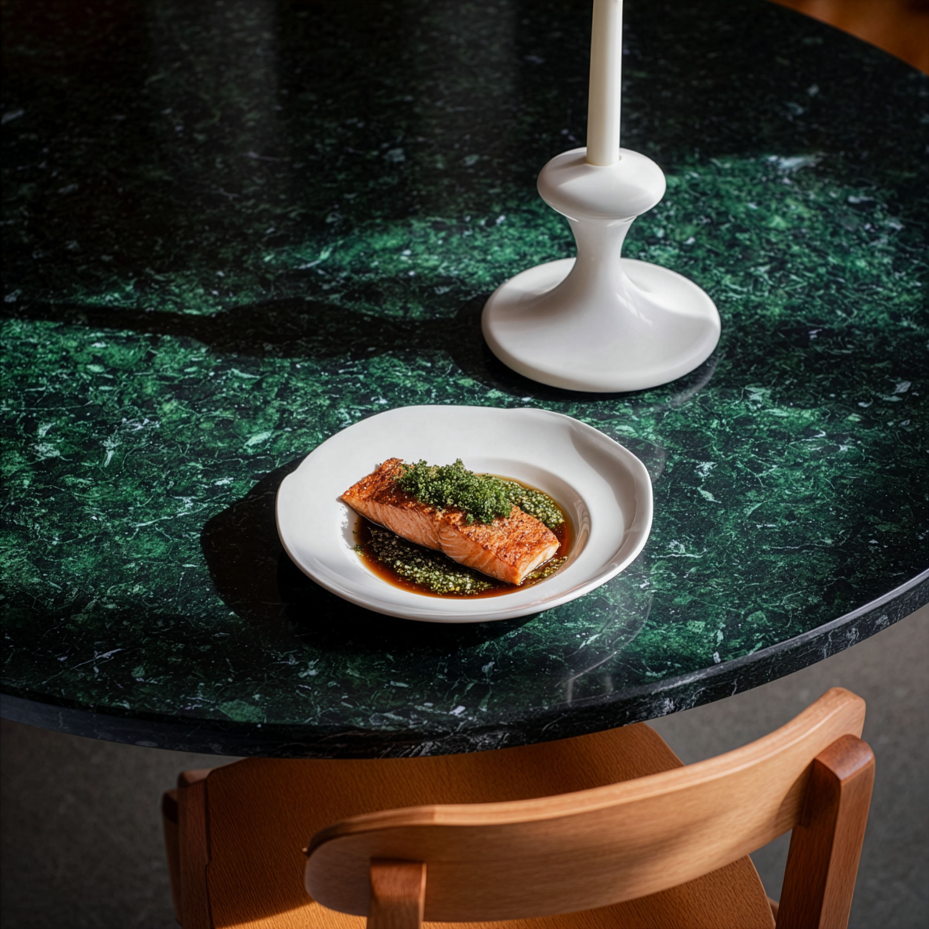 Plate with a seared salmon fillet topped with green herb garnish and sauce, served on a white dish placed on a dark green marble table beside a white candlestick.