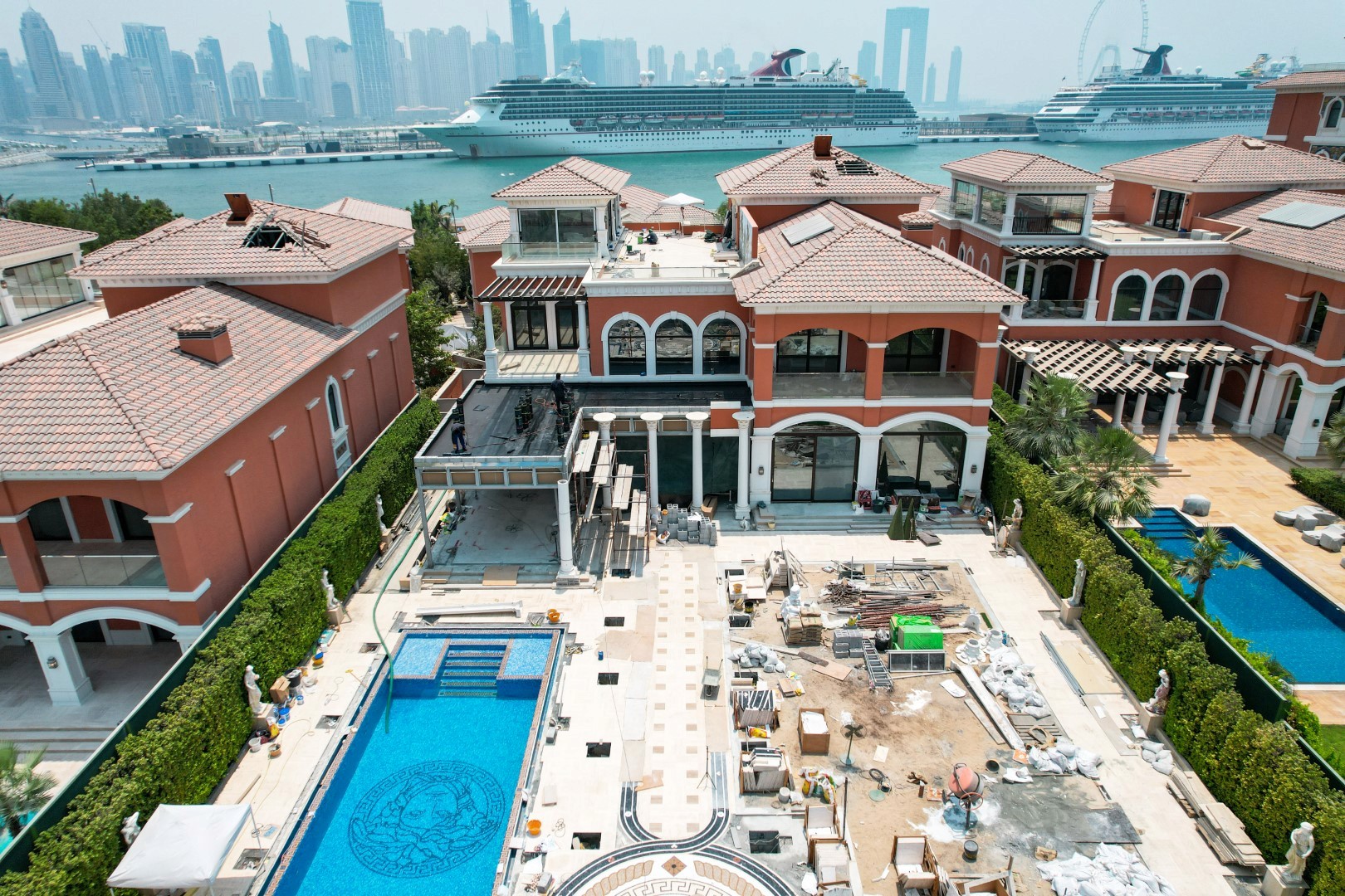 A luxurious waterfront villa under construction, featuring a partially built pool and surrounding landscaping, with a backdrop of towering city skyscrapers and two large cruise ships docked nearby.