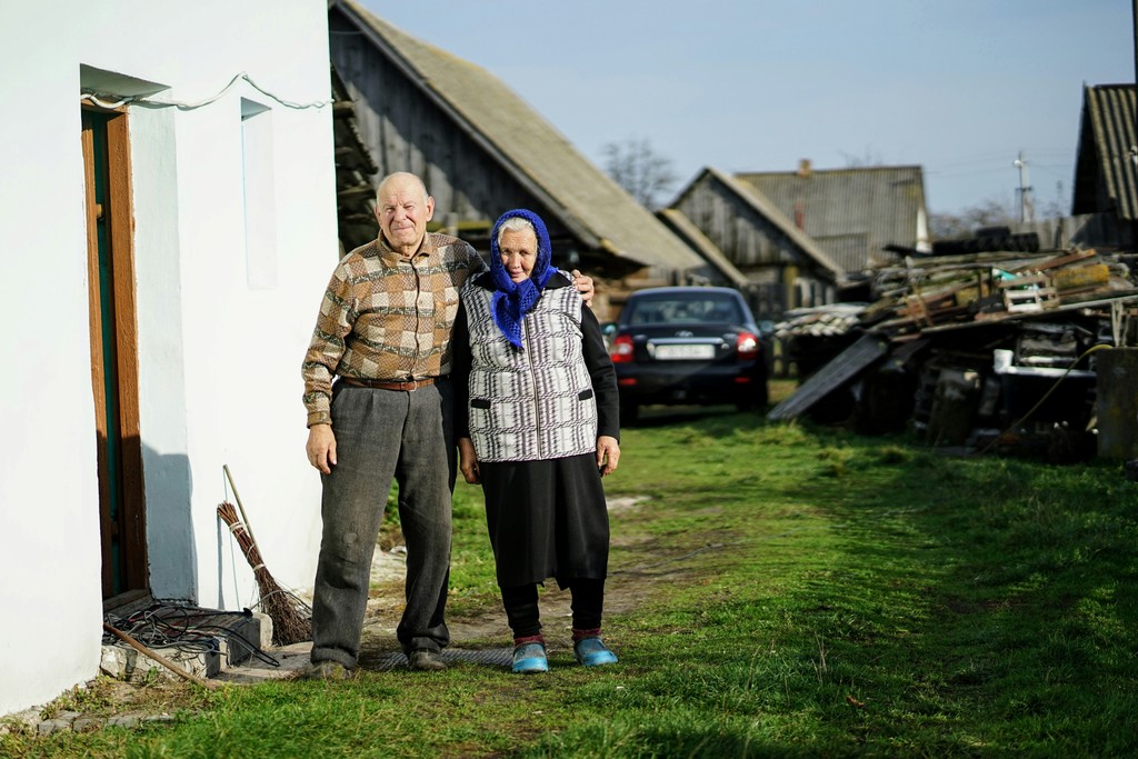 This heartwarming image captures an elderly couple standing outside their rural home, exuding a sense of warmth and resilience. The man, wearing a patterned shirt and gray pants, has his arm around the woman, who is dressed in a headscarf, checkered vest, and black skirt. The rustic setting features old wooden buildings, a parked car, and scattered farming equipment, reflecting a simple yet enduring lifestyle. The green grass and clear sky add a serene backdrop, enhancing the feeling of homely comfort and long-lasting companionship.