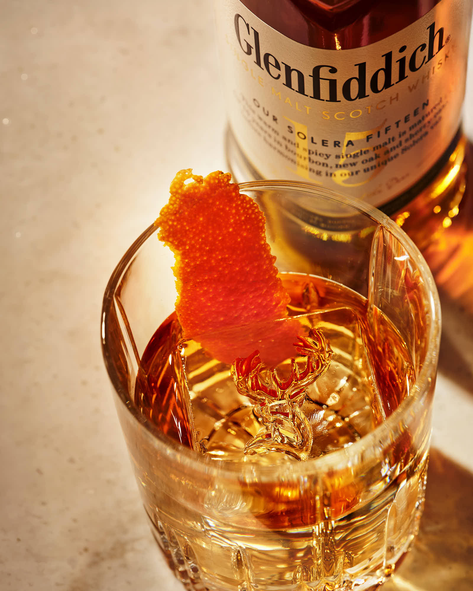 A glass of whiskey with an orange twist, alongside a bottle in a soft, blurred background.