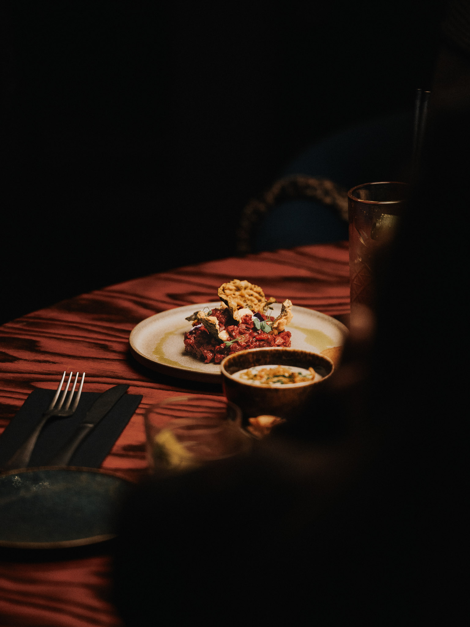 Wooden outdoor table set with multiple plates of food, including dips, salads, potatoes with cheese, and glasses of water, with hands reaching in to eat.