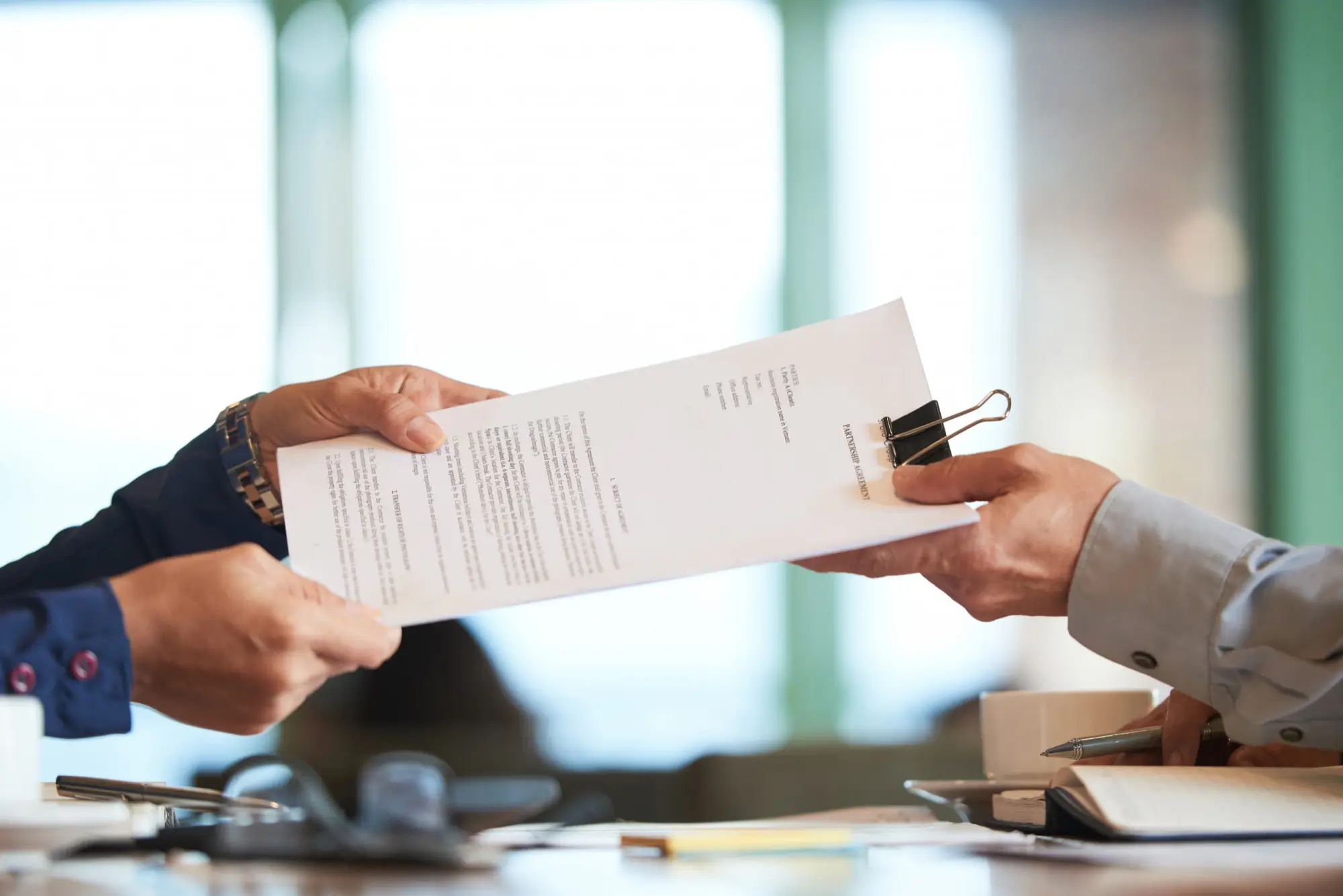 Two professionals exchanging a clipped contract document across a desk in an office setting.