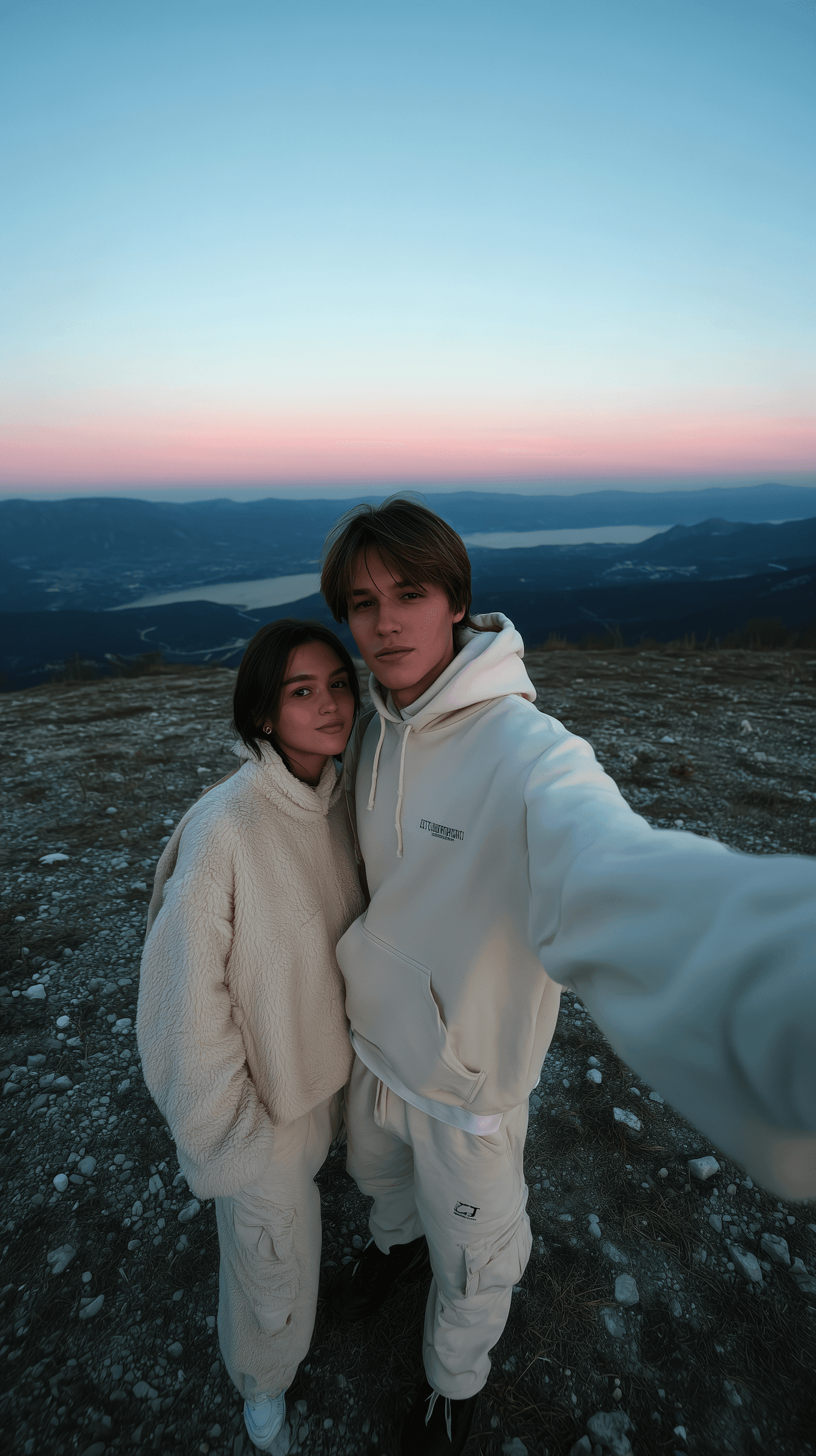 Two people in white jackets pose for a selfie against a scenic sunset backdrop with mountains.