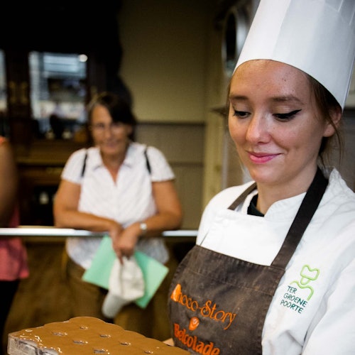 Egy fehér sapkát és kötényt viselő séf egy tálcát tart, miközben a háttérben egy nő figyel, zöld mappát tartva.