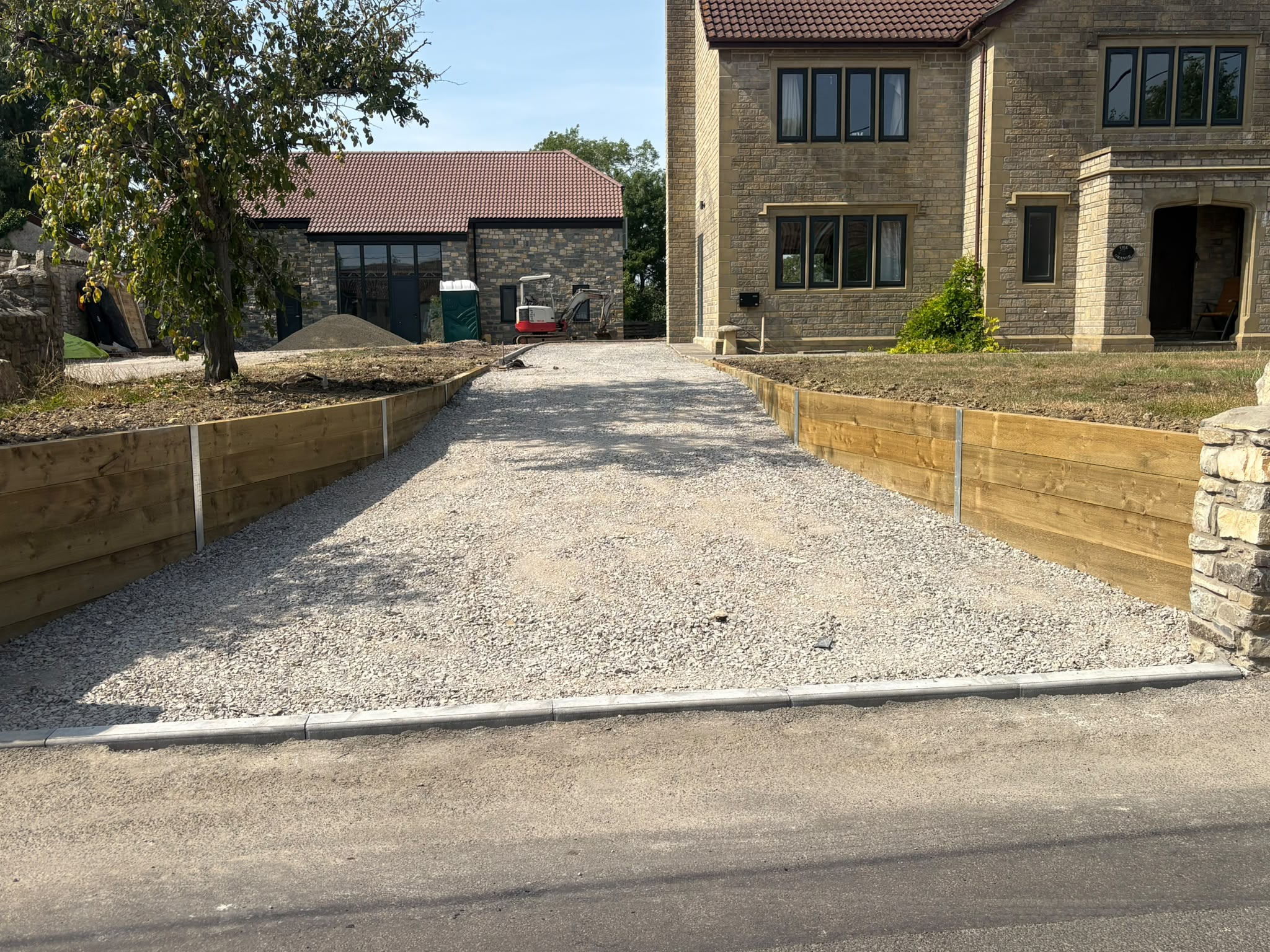 Gravel driveway leading to a stone house, bordered by timber retaining walls and a low stone entrance wall.