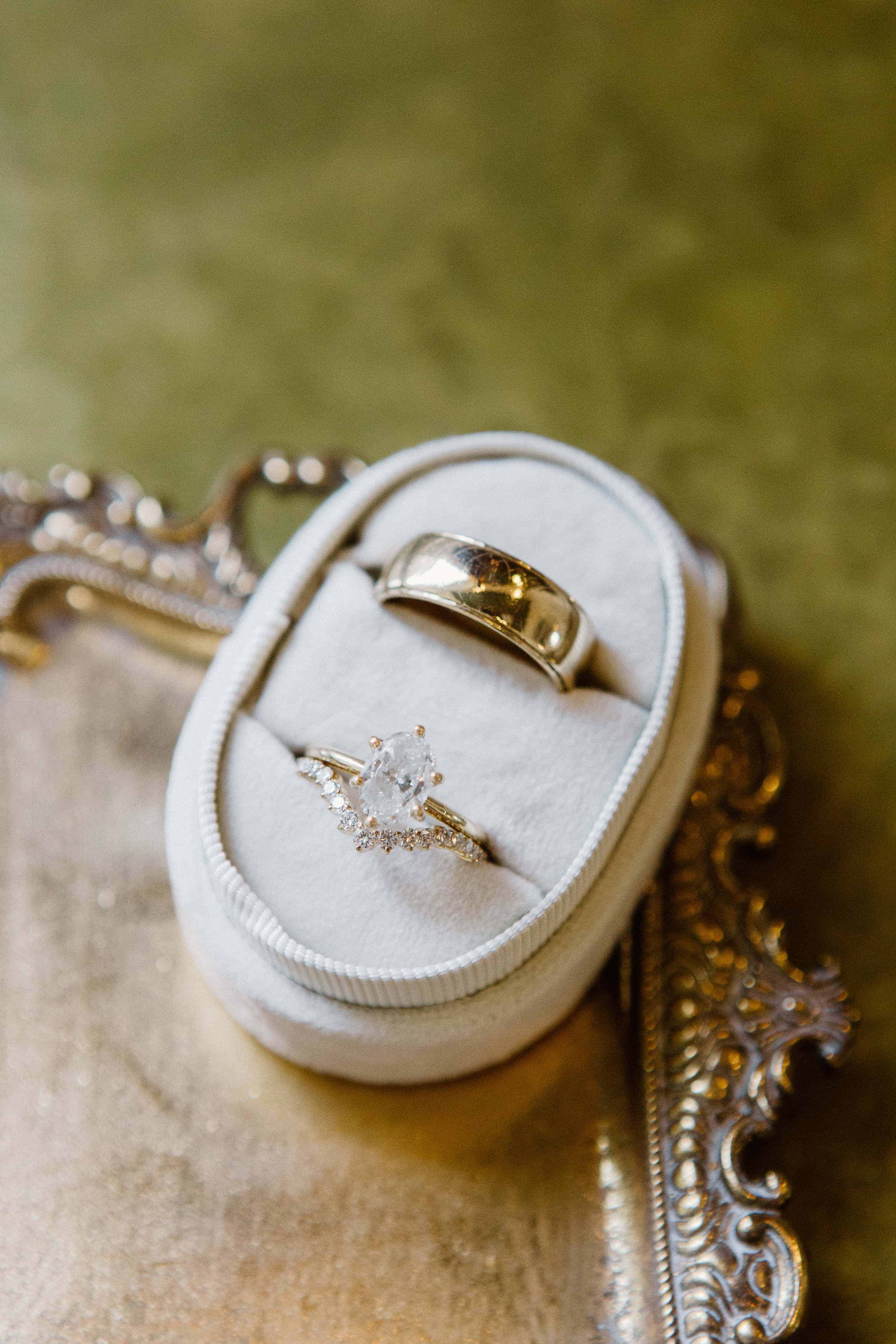 A close-up of a wedding ring set, with a diamond engagement ring and a solid gold wedding band, sitting in a velvet box on a decorative gold tray.
