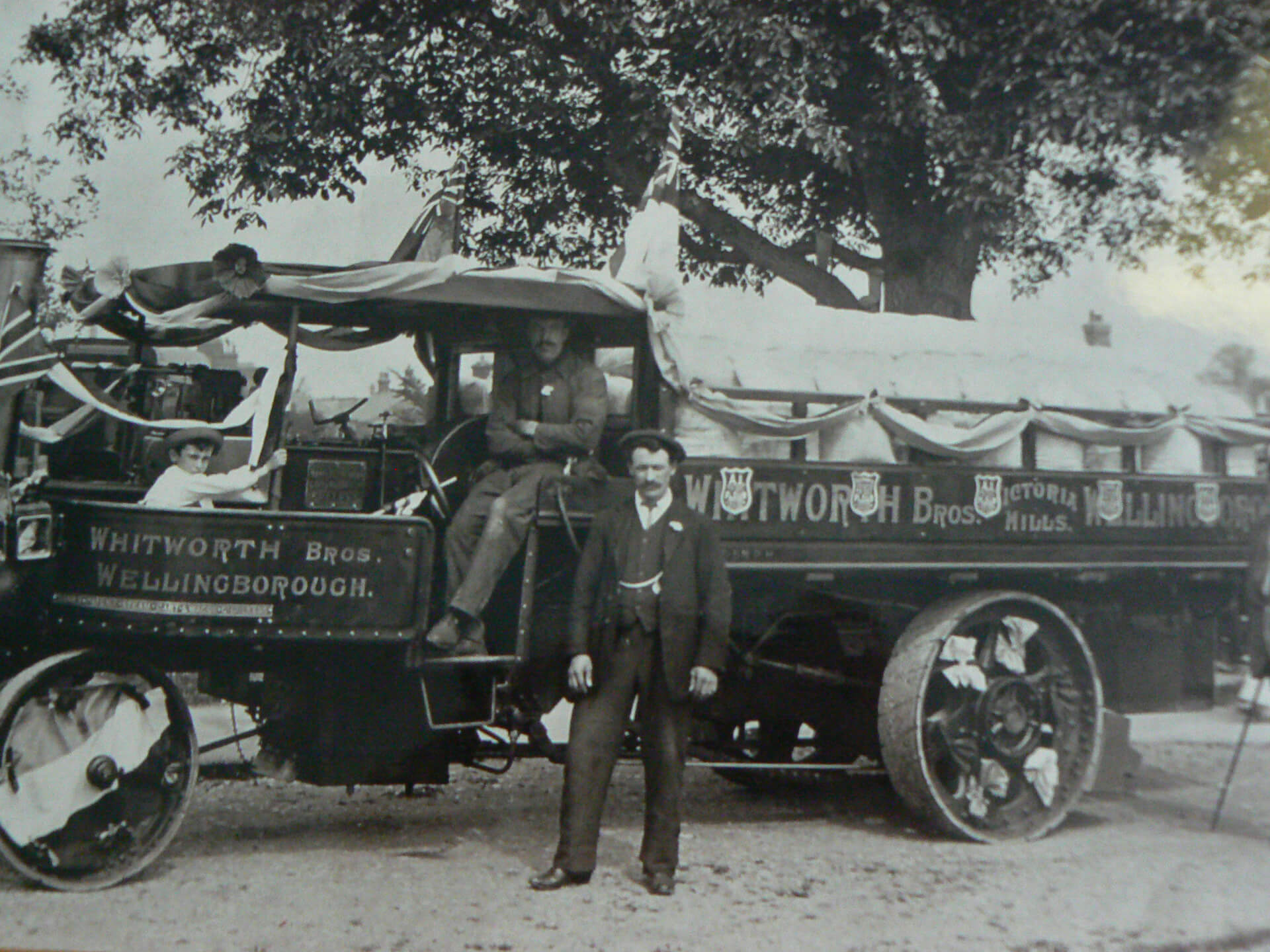 Historic photo of a man standing next to an old bus, with passengers visible inside, under a tree backdrop.