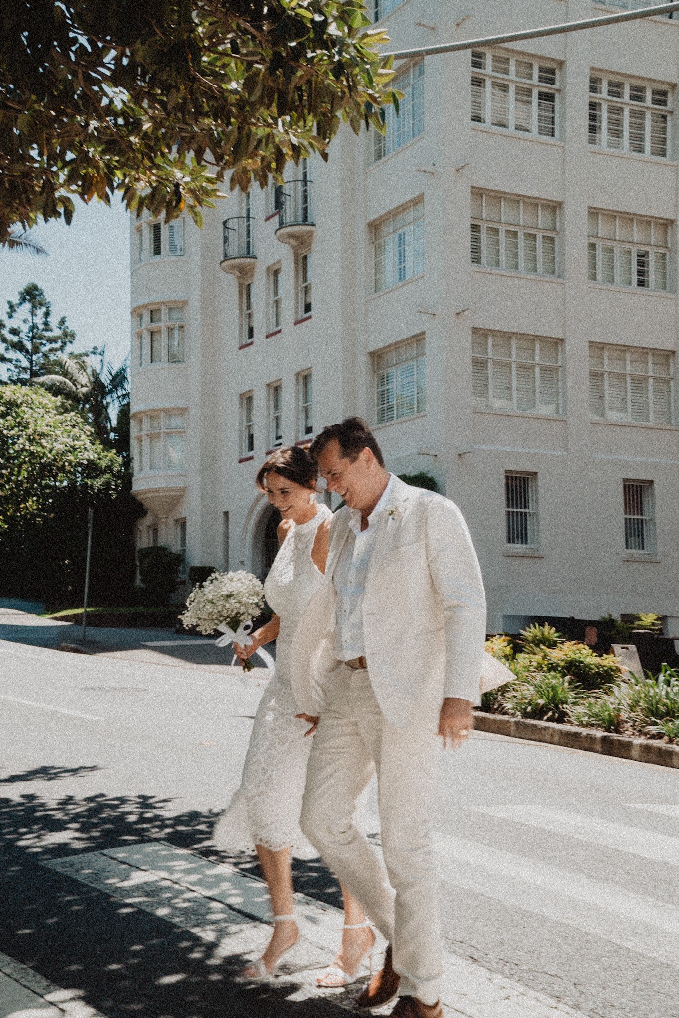 Bride and groom in white walking across a city street with a bouquet.