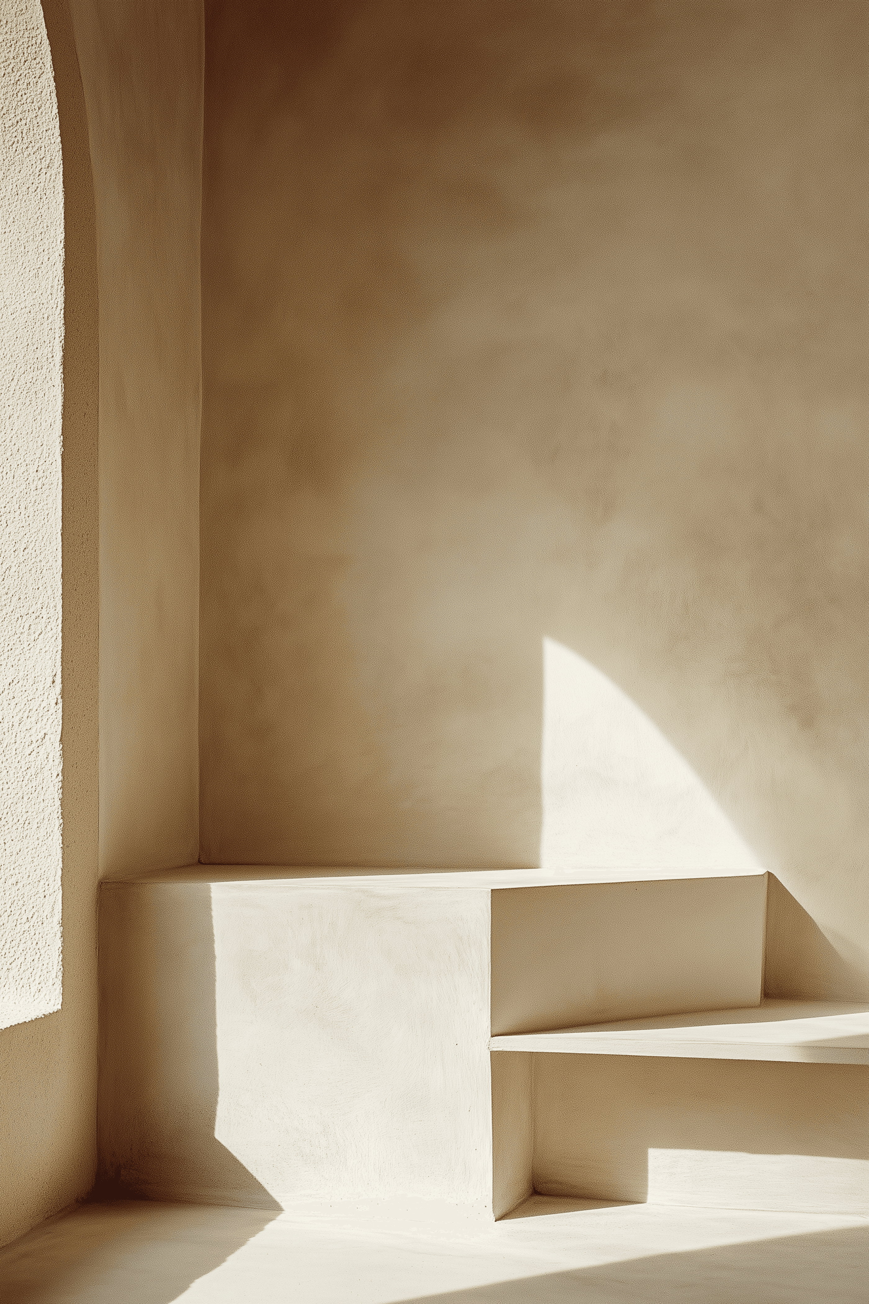 A single low plaster step built into the corner of an empty beige room, lit by a curved slice of sunlight from an arched window