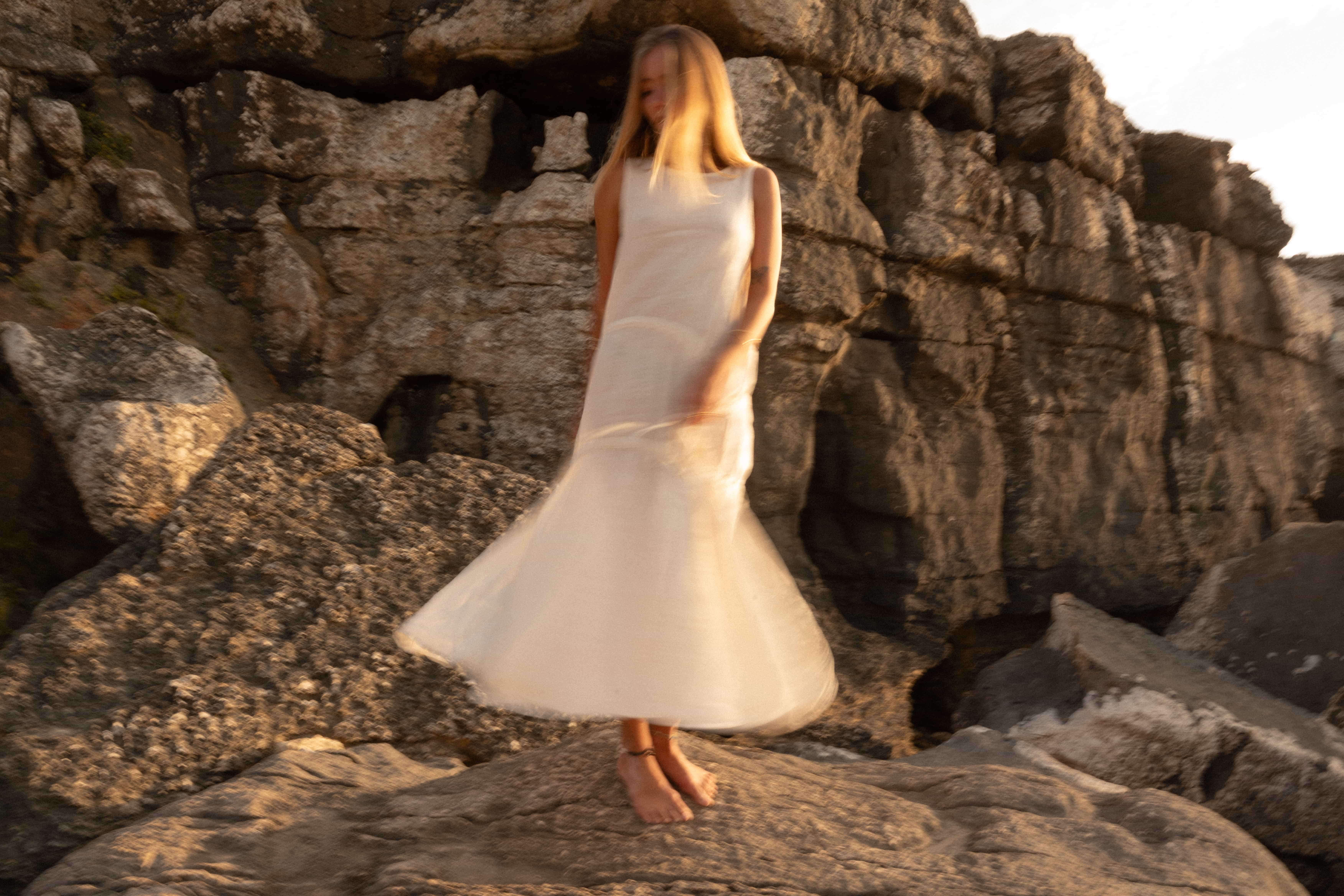 Woman twirling in a flowing white dress on coastal rocks at sunset