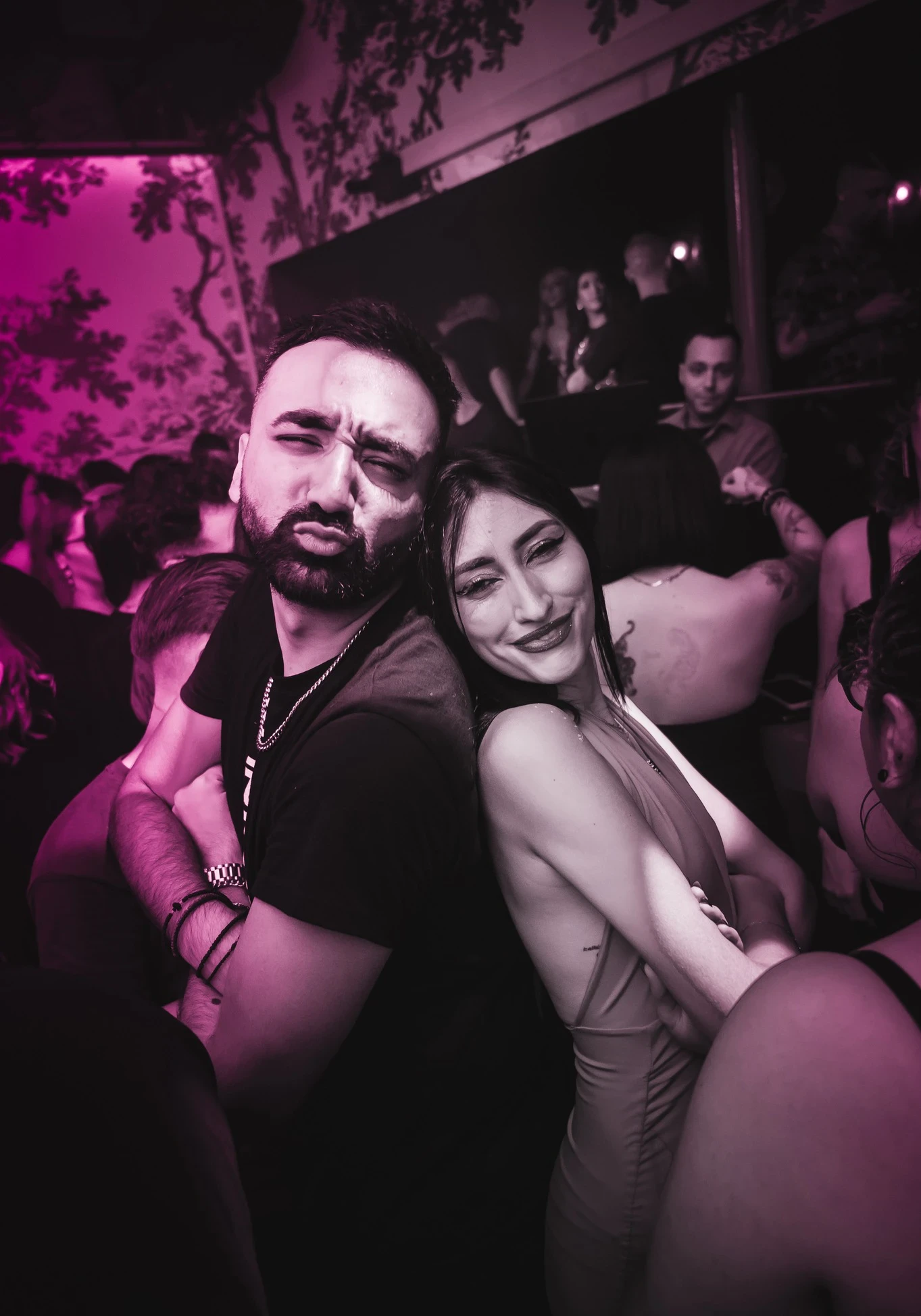 Liakakis and a woman pose back-to-back in a crowded club. The man makes a pouty face, and the woman smiles at the camera.