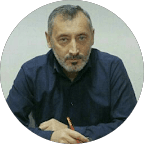 Blonde woman in a black long-sleeve and apron, arms crossed—minimalist studio portrait on a beige background.