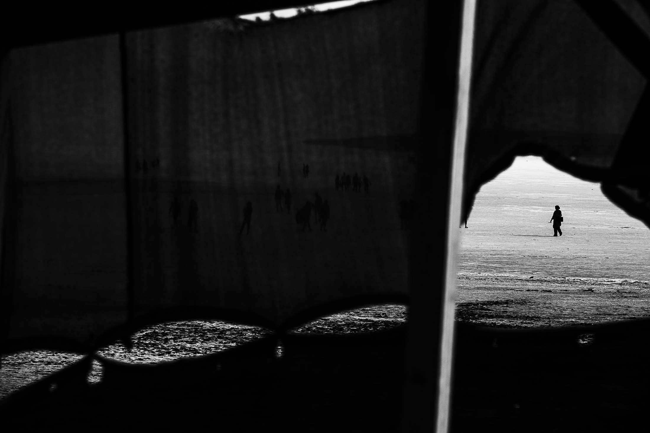 Small child walking alone across an open beach, seen through dark foreground framing elements.