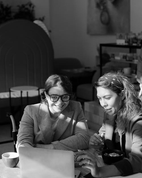 Two women sitting together in a cozy, well-lit workspace, smiling and collaborating over a laptop, with a coffee cup and a bowl on the table.