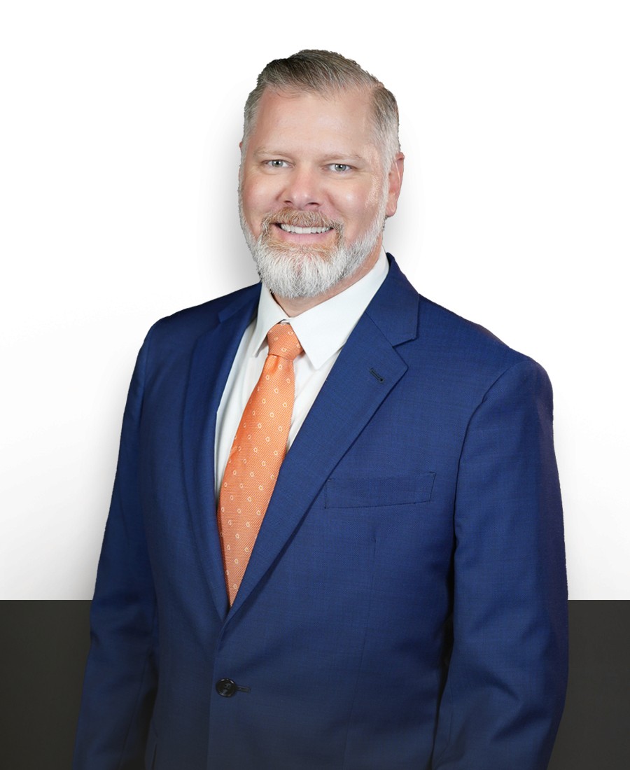 corporate headshot of a male with light skin and grey hair, mustache, and beard, wearing a royal blue suit with white shirt and orange, white-spotted tie. He is smiling casually. 