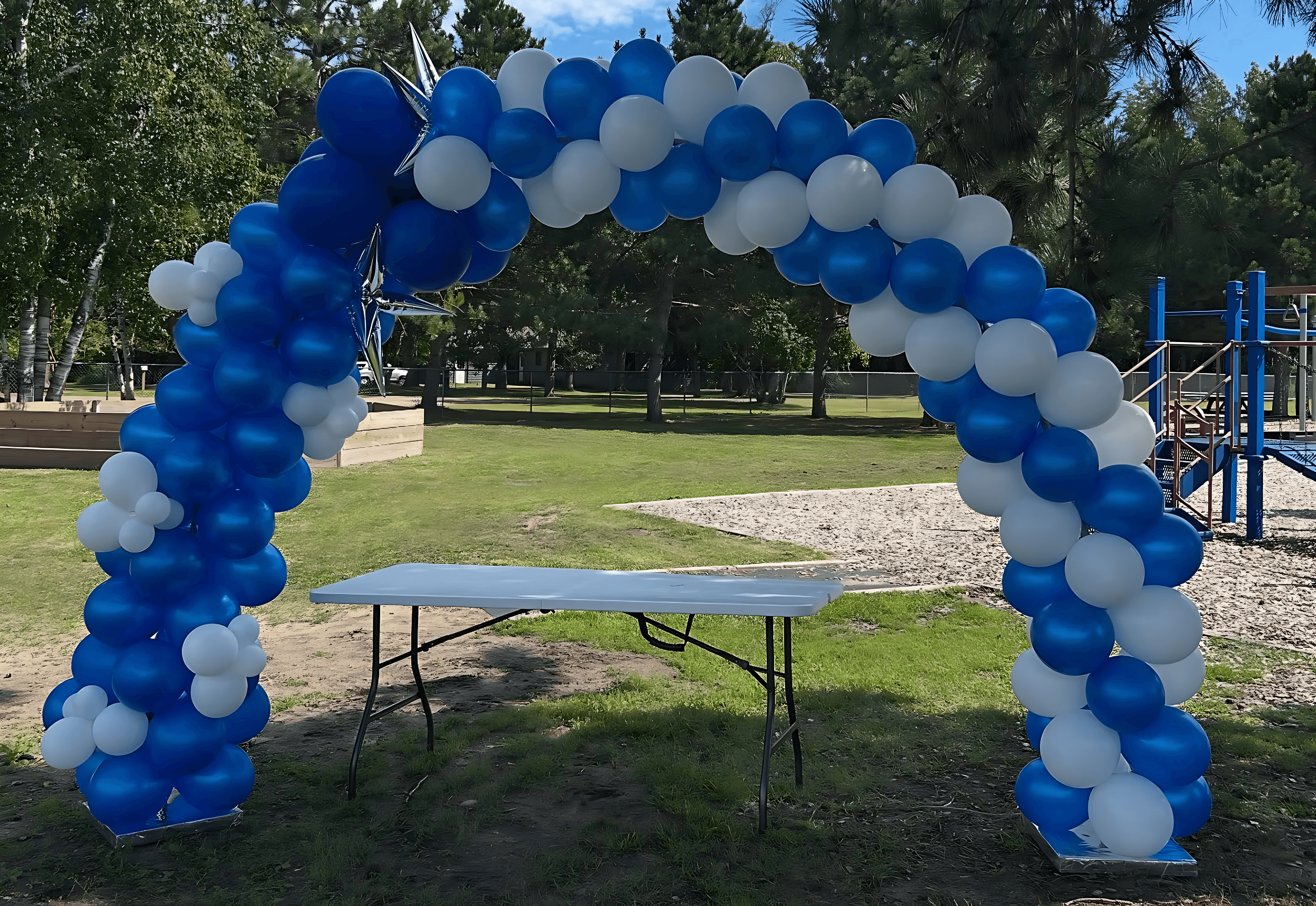 A festive arch made of alternating blue and white balloons stands over a white folding table on a grassy area in a park, with trees and a playground visible in the background.
