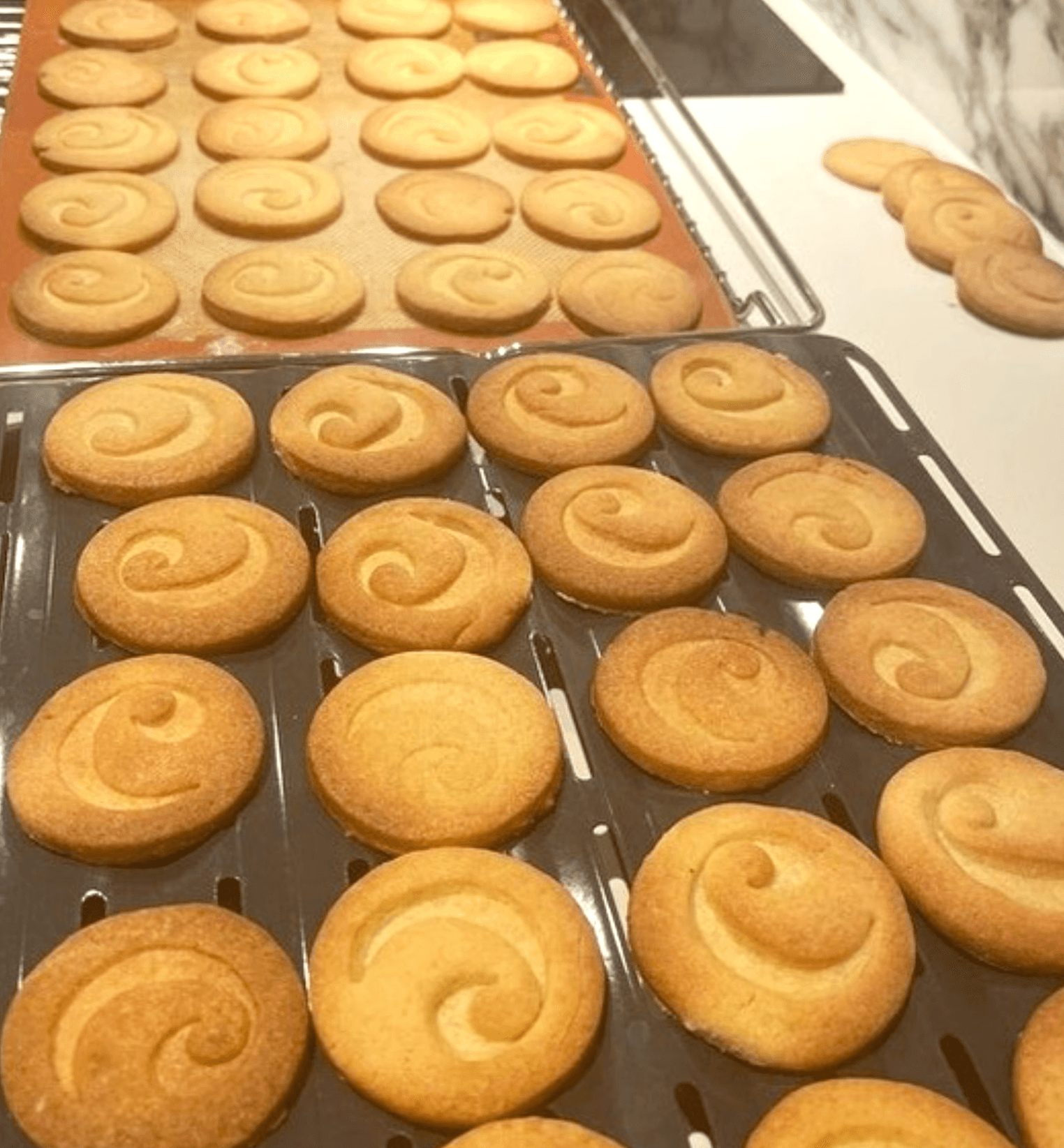Multiple trays of golden-brown swirl cookies cooling on wire racks in a kitchen setting, arranged in neat rows after baking, with Carefree logo on each.