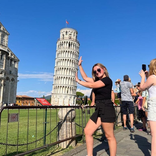 Žena vtipně pózuje, jako by držela Šikmou věž v Pise, obklopená dalšími turisty během slunečného dne.