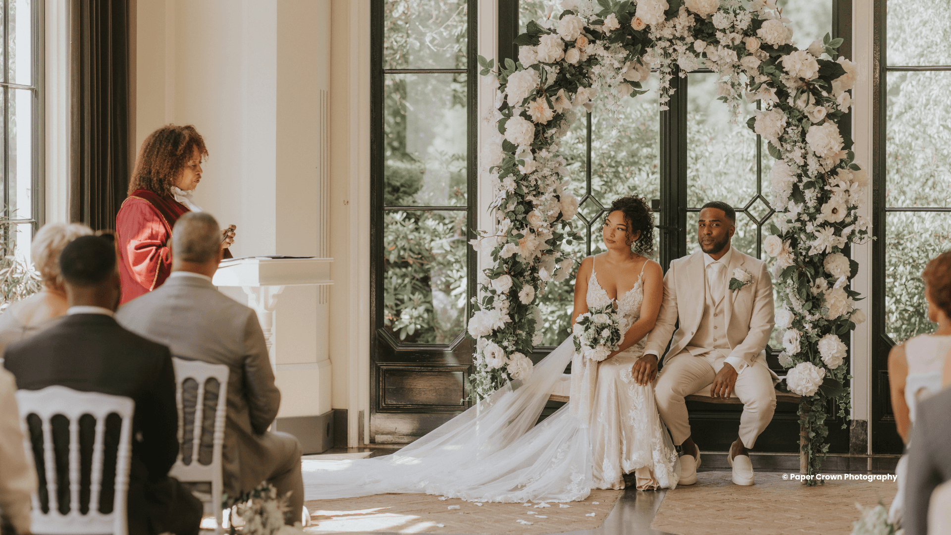 Een zelfstandig trouwambtenaar leidt een huwelijksceremonie terwijl het bruidspaar voor een bloemenboog zit, omringd door gasten tijdens een romantische trouwdag.