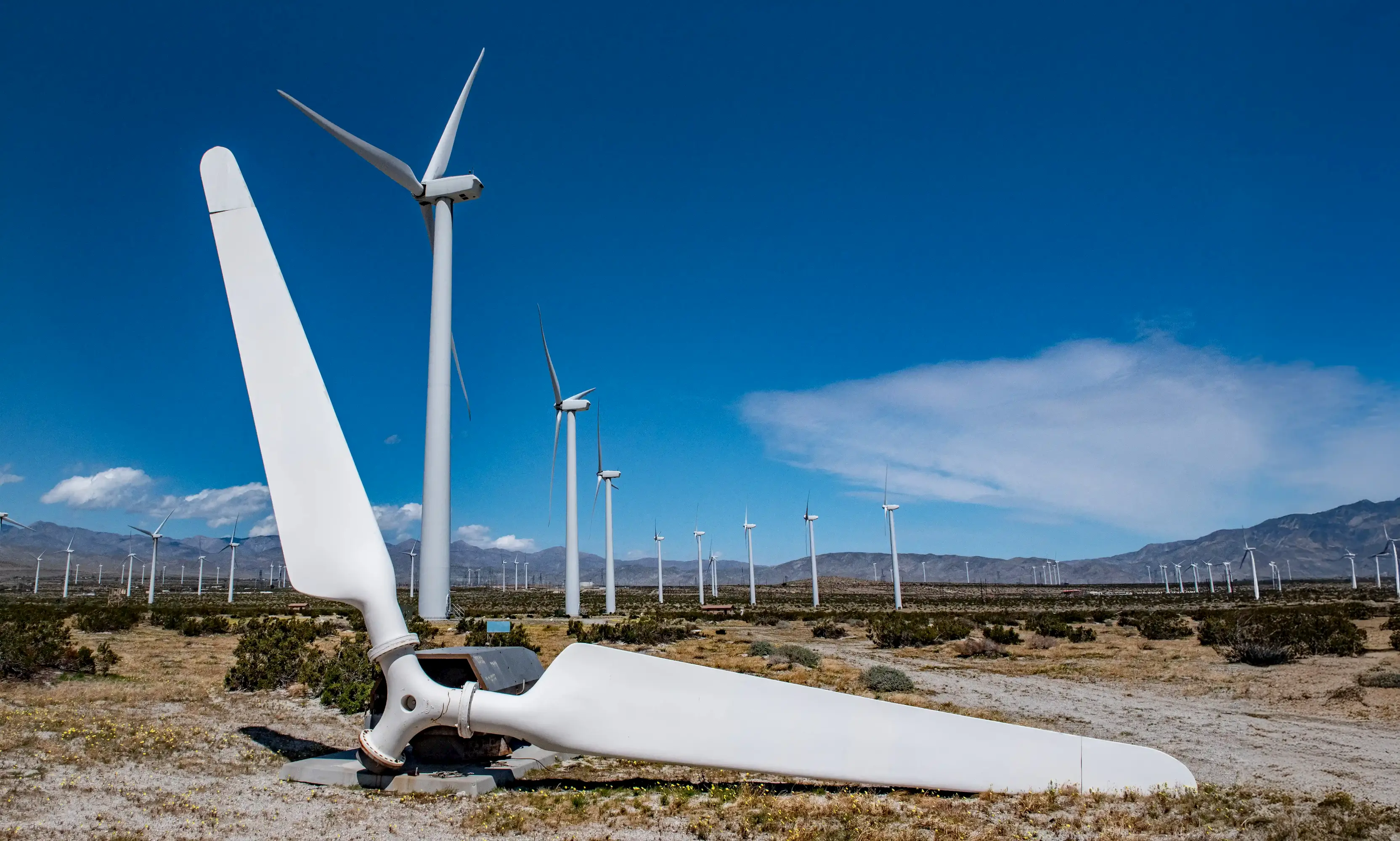 A large broken wind turbine blade lies on the ground in front of several intact wind turbines in a desert wind farm, with clear blue skies and mountains in the background.