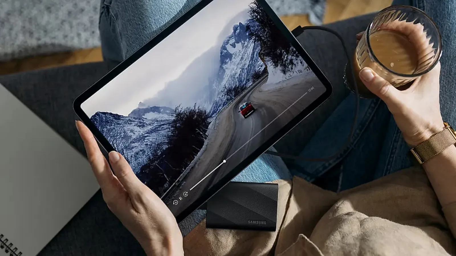 A person is sitting on a couch holding a glass of beverage in one hand and a tablet in the other, watching a scenic video of a car driving on a snowy mountain road, with a Samsung external SSD visible nearby.