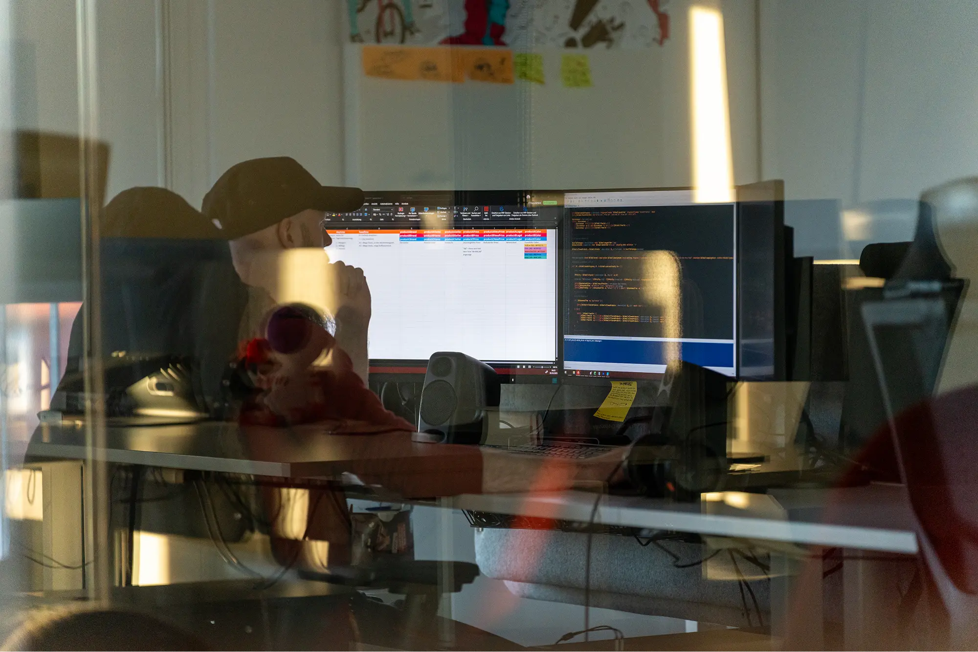 Ein fokussierter Entwickler arbeitet konzentriert an mehreren Bildschirmen mit Code- und Datenzeilen, fotografiert durch die Spiegelung einer hellen Büroscheibe.
