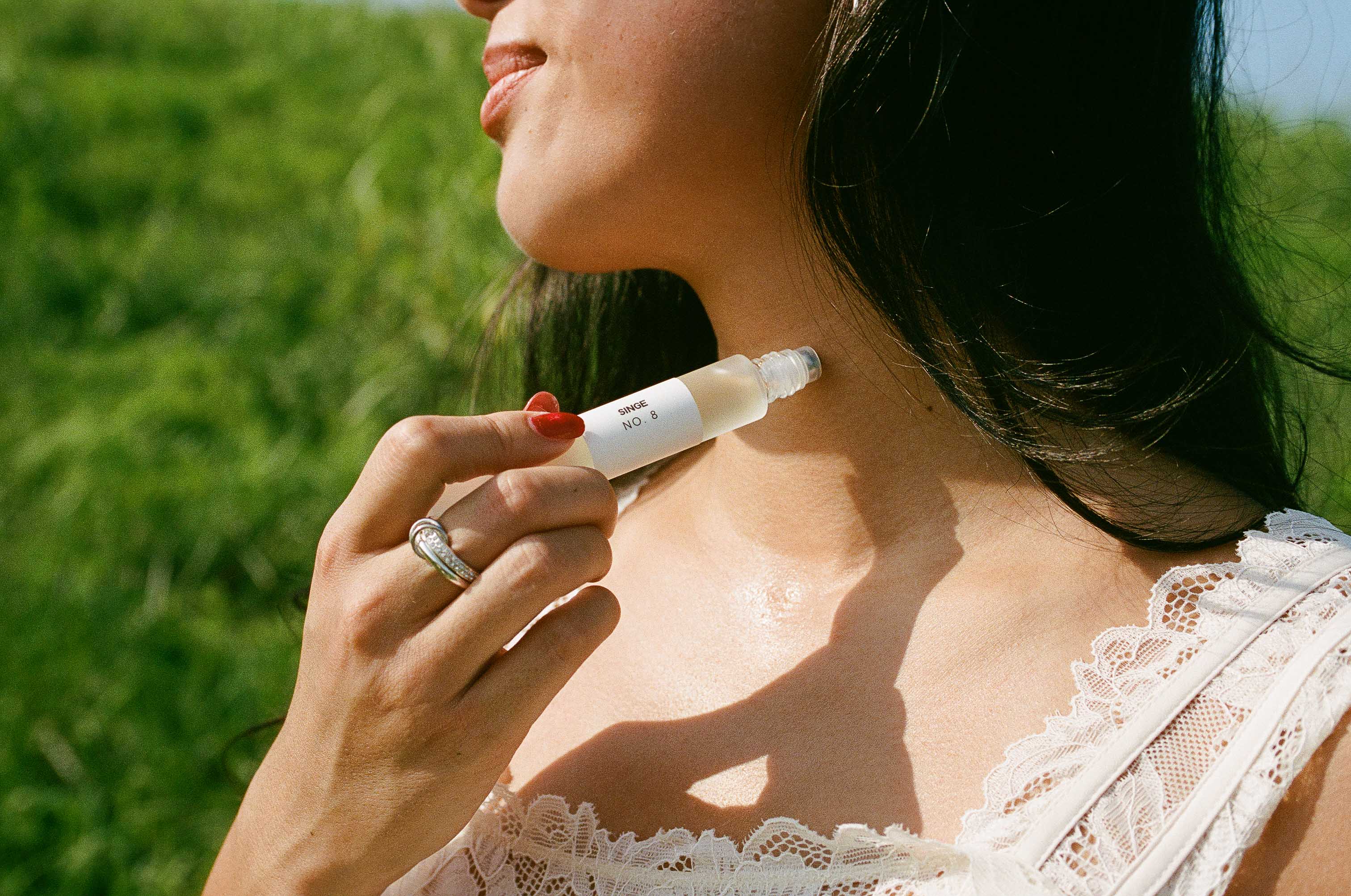 A close-up lifestyle shot of a woman outdoors in a grassy field, applying SINGE No. 8 rollerball perfume.