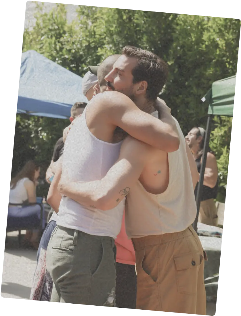 Two queer people hugging each other’s shoulder in a quiet, affectionate moment.