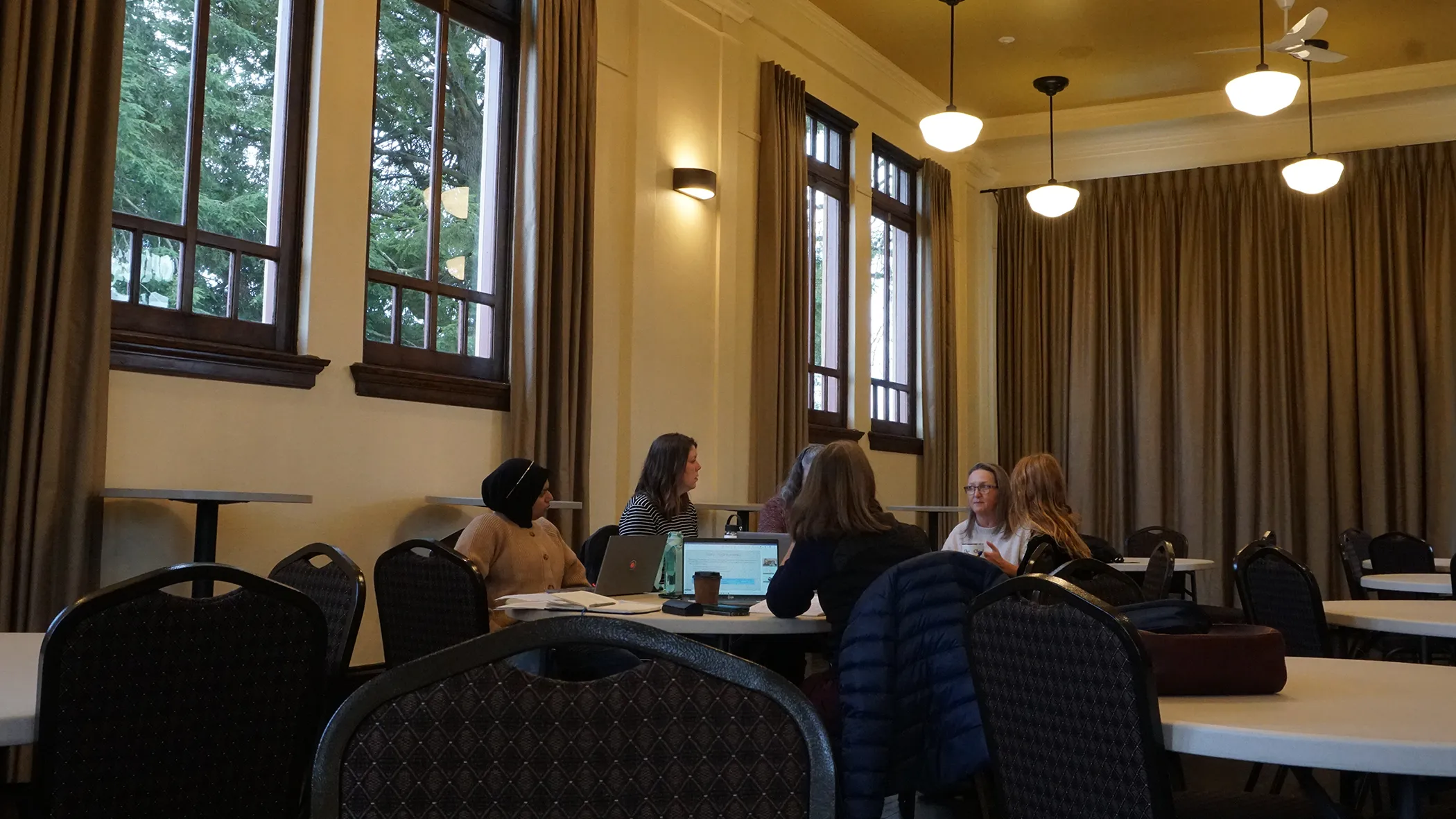 A meeting with 6 people around a round table with laptops out. They are in a larger room with a high ceiling, there are similar tables set up around them, and the lighting is dim and warm.