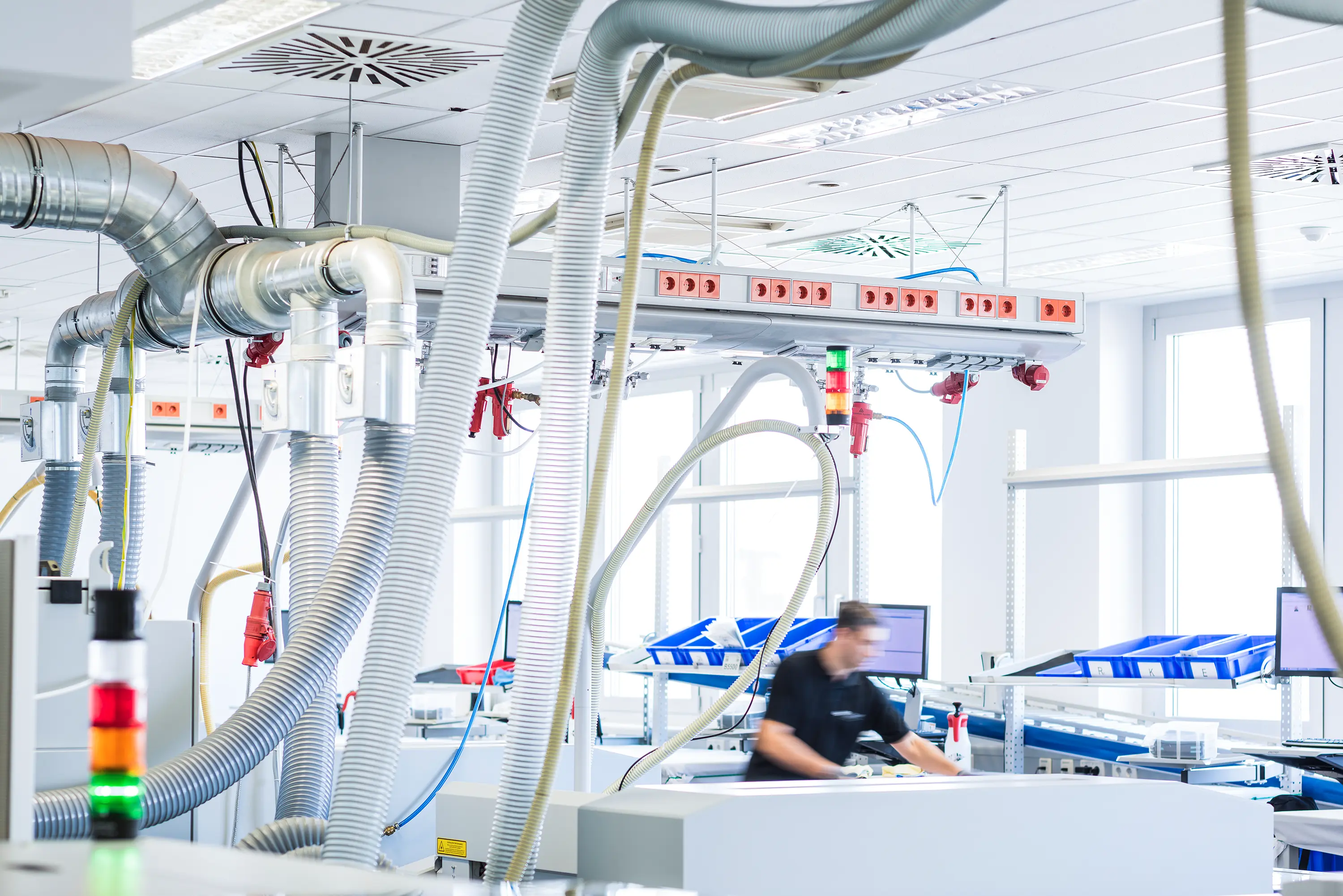 Blick in eine helle Produktionshalle mit technischen Absaugschläuchen und einem Mitarbeiter an einer Arbeitsstation