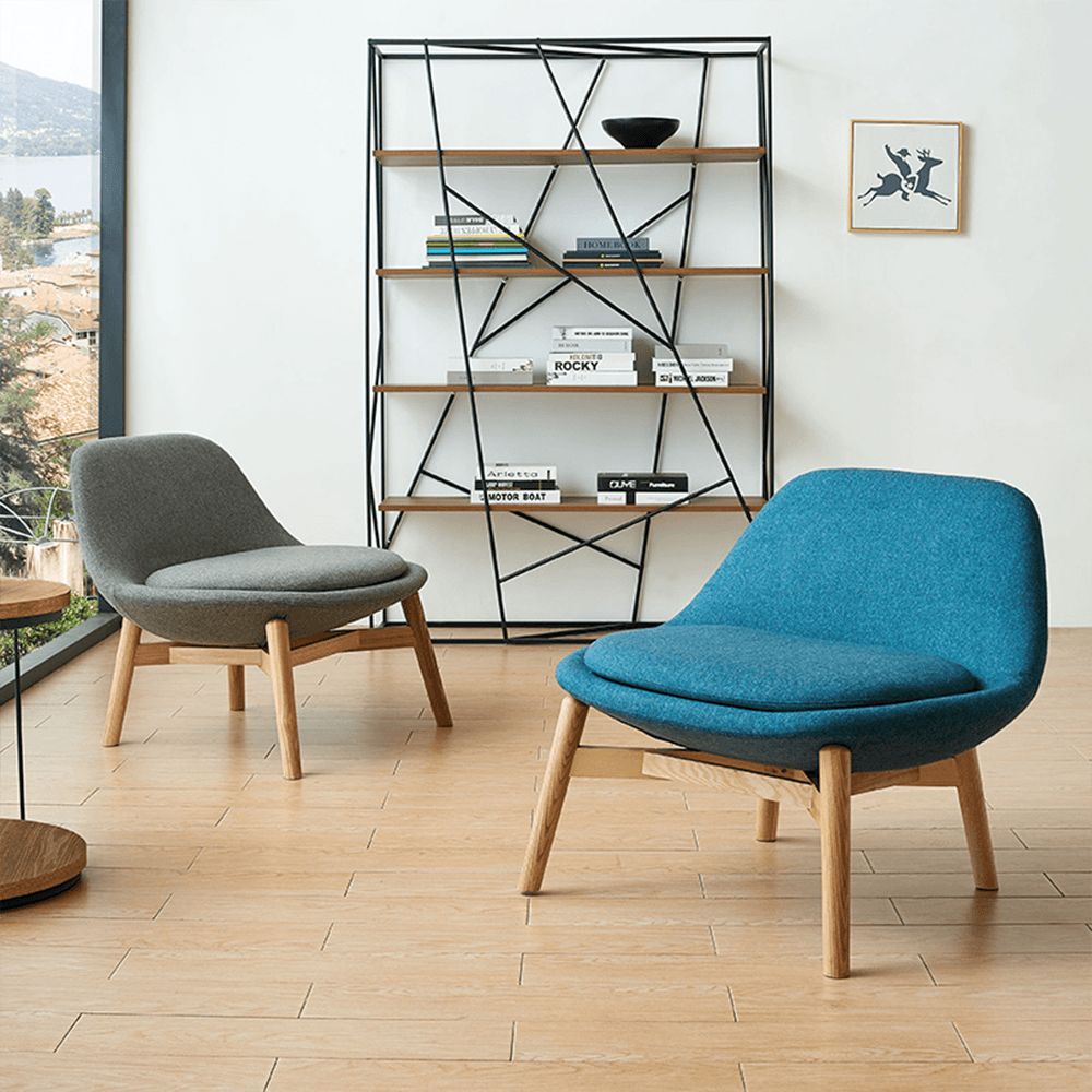 Pair of modern curved lounge chairs in gray and teal blue fabric with oak legs, beside geometric black metal bookshelf in bright lakeside living room – mid-century modern furniture setup