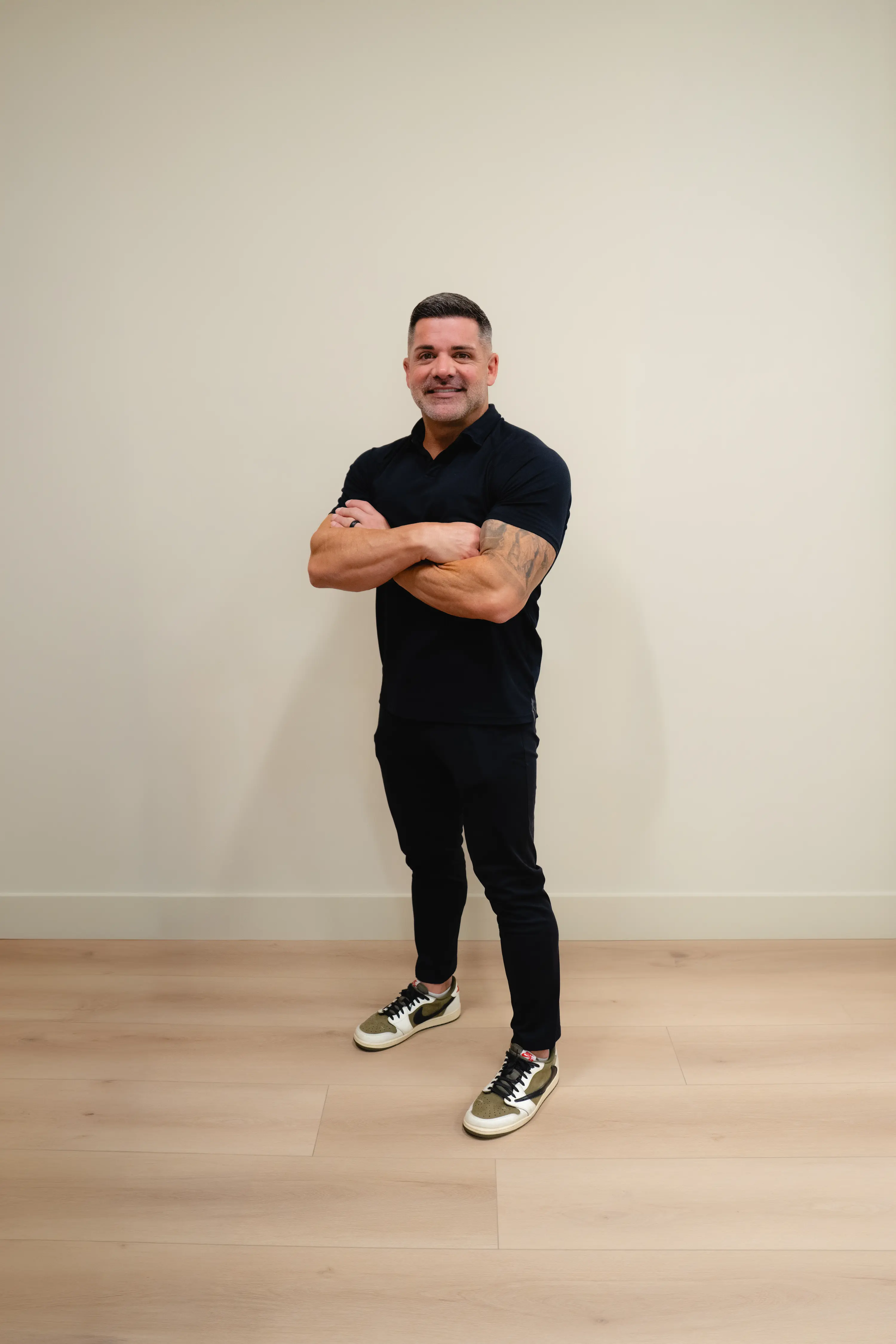 Man in a black polo standing with arms crossed against a white wall