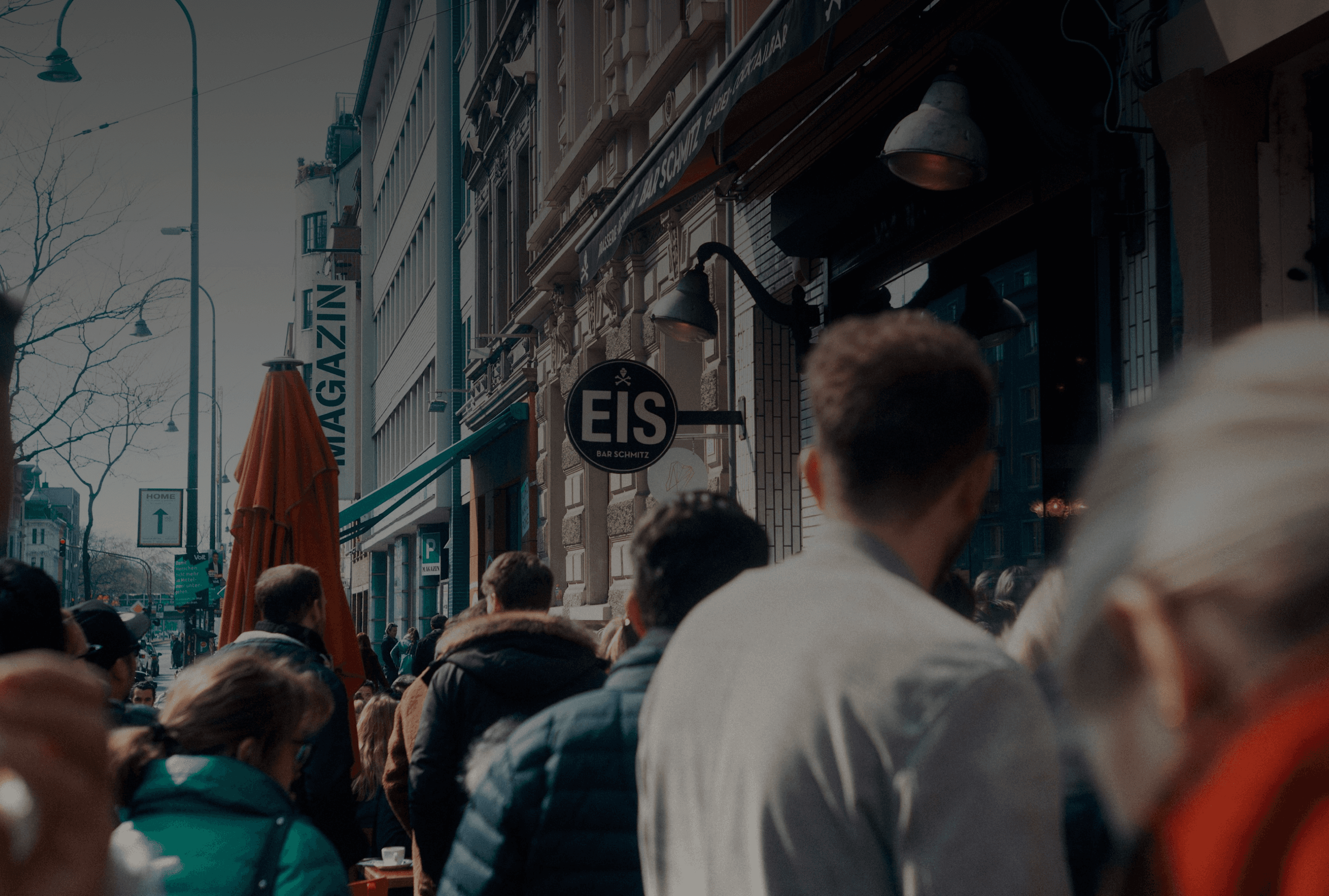 Una escena de calle concurrida con personas reunidas afuera de una tienda o café. El letrero sobre la tienda dice "EIS", lo que sugiere que podría ser una heladería o una tienda de postres. El entorno parece una zona urbana abarrotada.