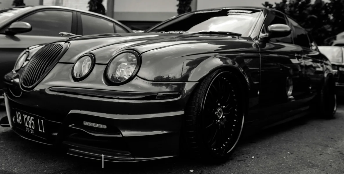 Un cliché en noir et blanc d’une Jaguar profondément modifiée, posée très bas sur de larges jantes sombres. L’ambiance contrastée souligne le style agressif de la préparation et met en valeur les courbes classiques du modèle.