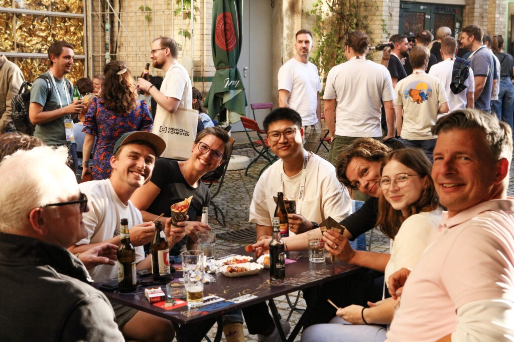 Gruppe von Menschen sitzt an einem Tisch im Freien und hält Getränke in der Hand, im Hintergrund weitere Personen in lockerer Atmosphäre vor einem Gebäude