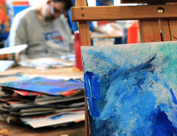A wooden easel holds a canvas with blue and white brushstrokes. The background is out of focus, showing a person wearing a light gray sweatshirt sitting at a table with stacks of papers or art supplies. The person’s face is blurred.