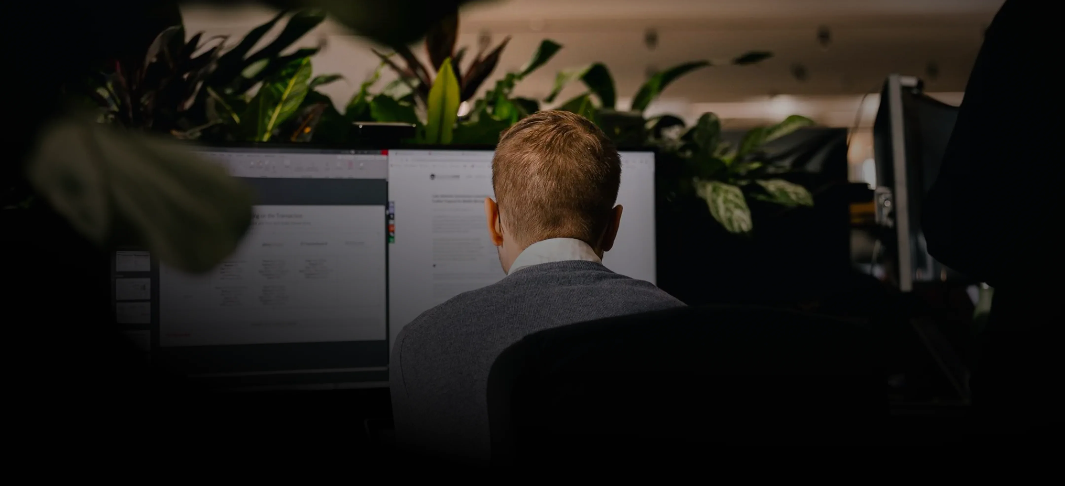A professional with light brown hair, wearing a gray sweater, works intently at a computer screen in a dimly lit office.
