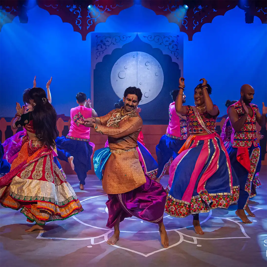 A group of dancers in vibrant traditional Indian attire perform energetically on stage, with blue and purple lighting and a large moon backdrop behind them.