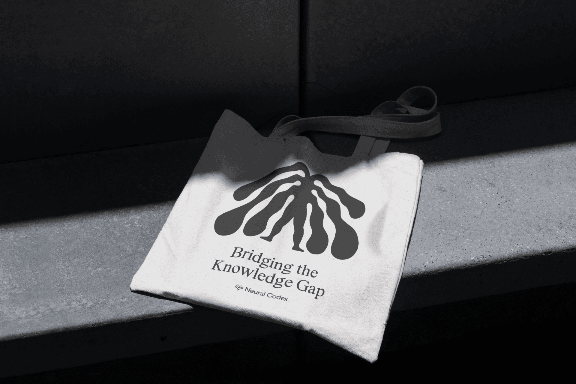 Cream Neural Codex tote bag on a dark bench in sunlight, printed with a grey abstract figure and “Bridging the Knowledge Gap”