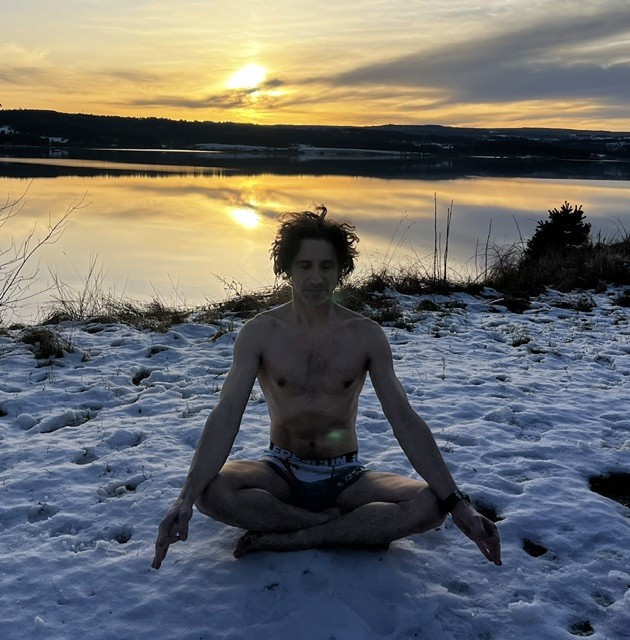Mathias méditant dans la neige près d’un lac, en sous-vêtements, avec les couleurs du lever du soleil se reflétant sur l’eau.