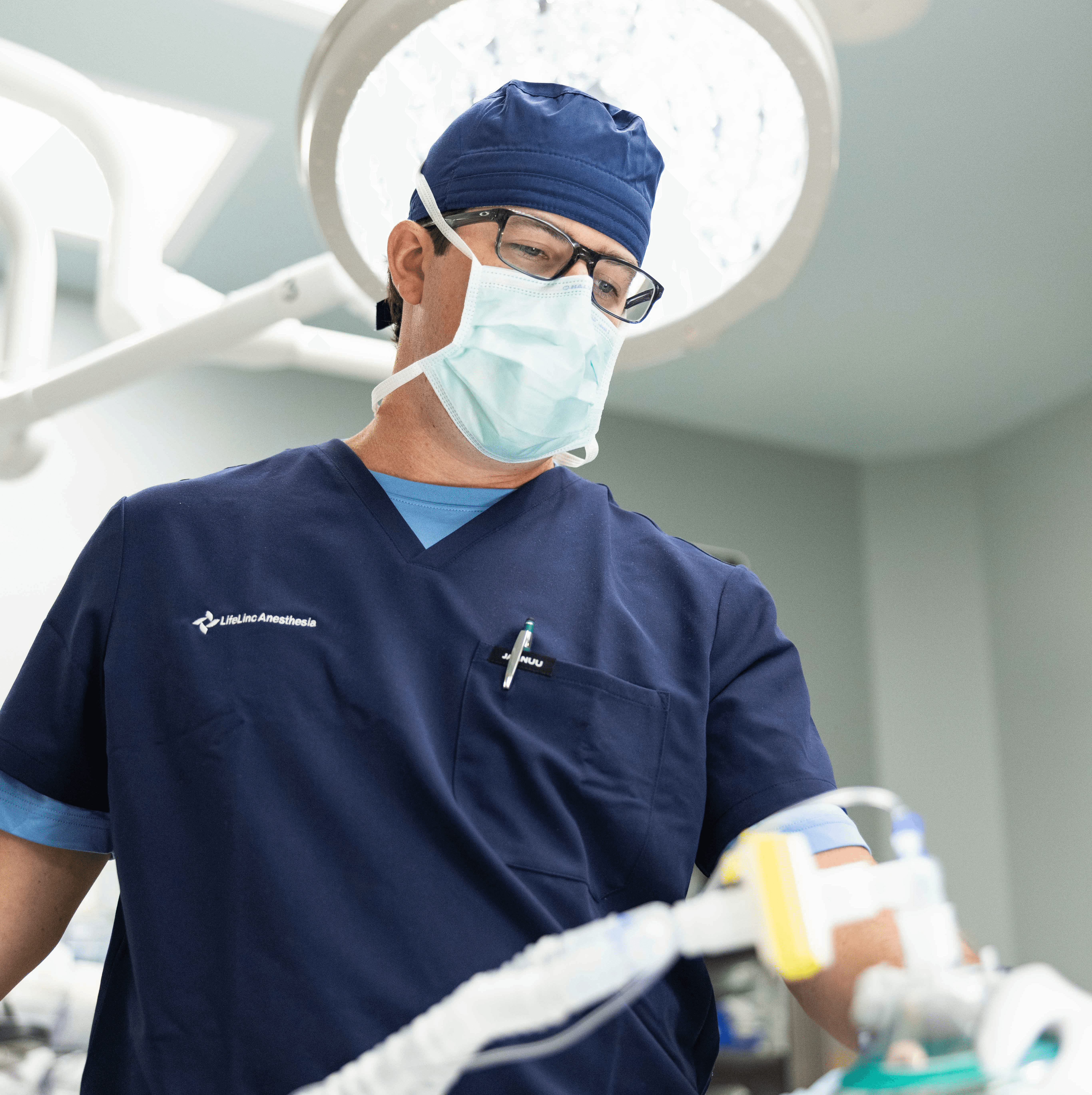A LifeLinc CRNA in navy scrubs, surgical cap, and mask focuses on anesthesia equipment during a procedure in an operating room, representing full-time employment opportunities